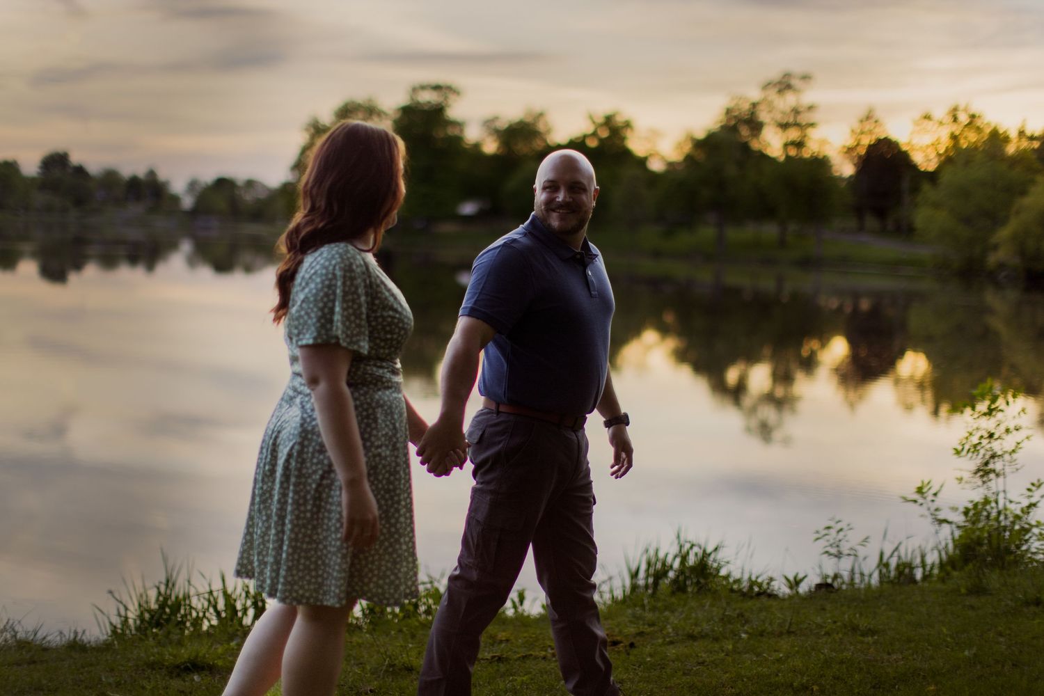A couple stands near a lake at sunset casting silhouettes against the golden sky and reflective water.