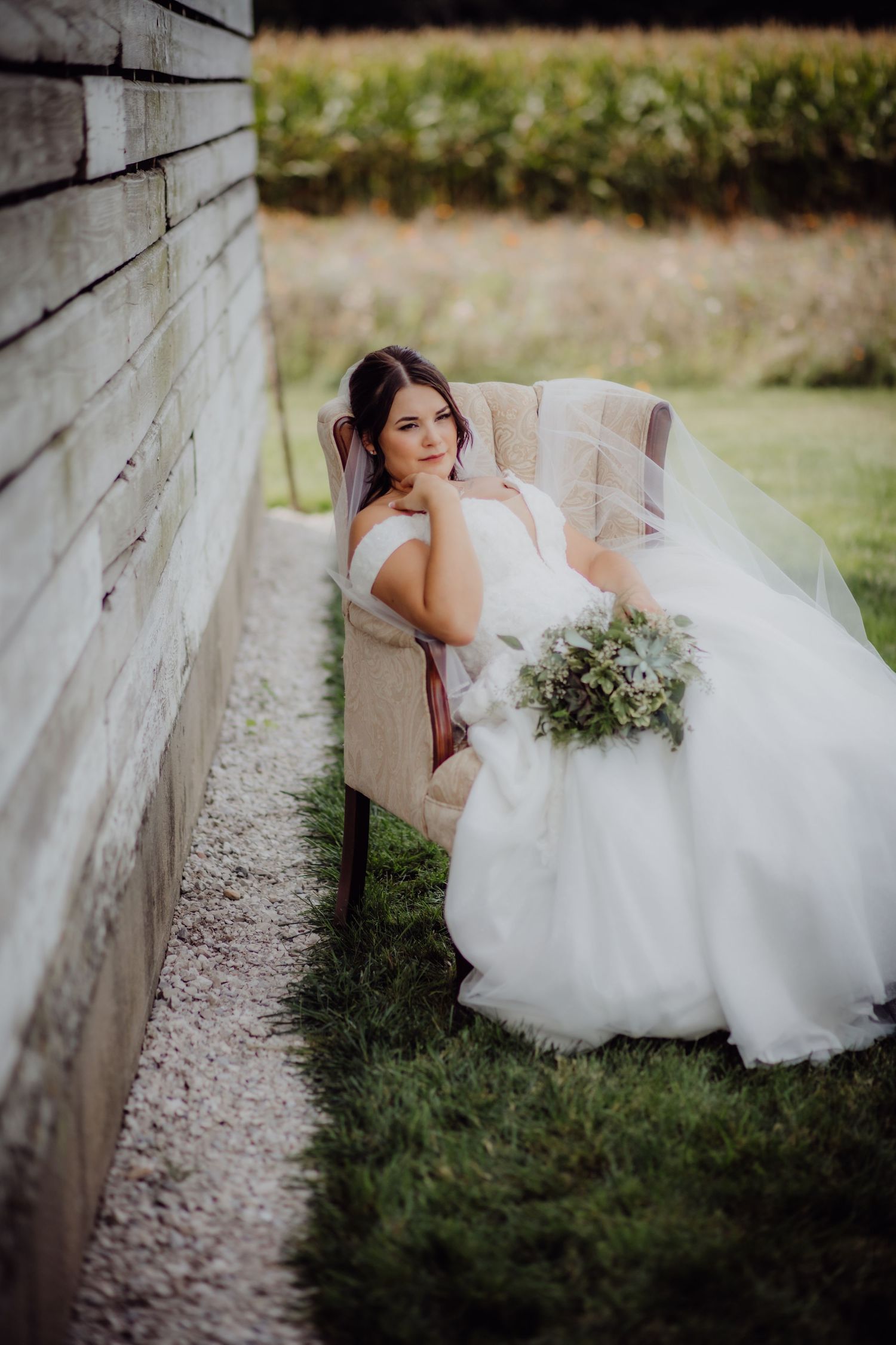Bride in white gown sits with bouquet against rustic wooden wall in outdoor setting.