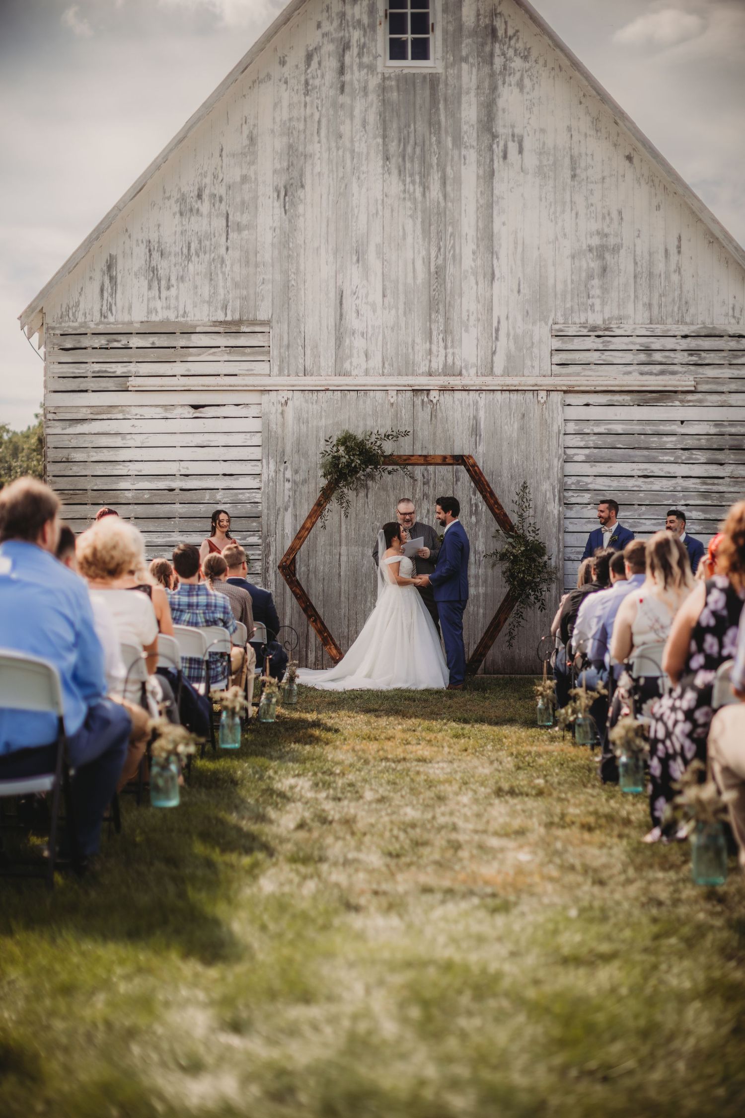Wedding celebration unfolds in front of a weathered white barn with guests gathered on the lawn.