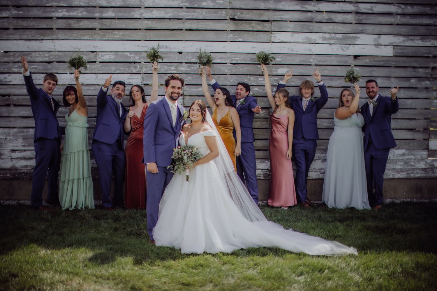 Bridal party dressed in multi-colored attire gathers against weathered wood siding.