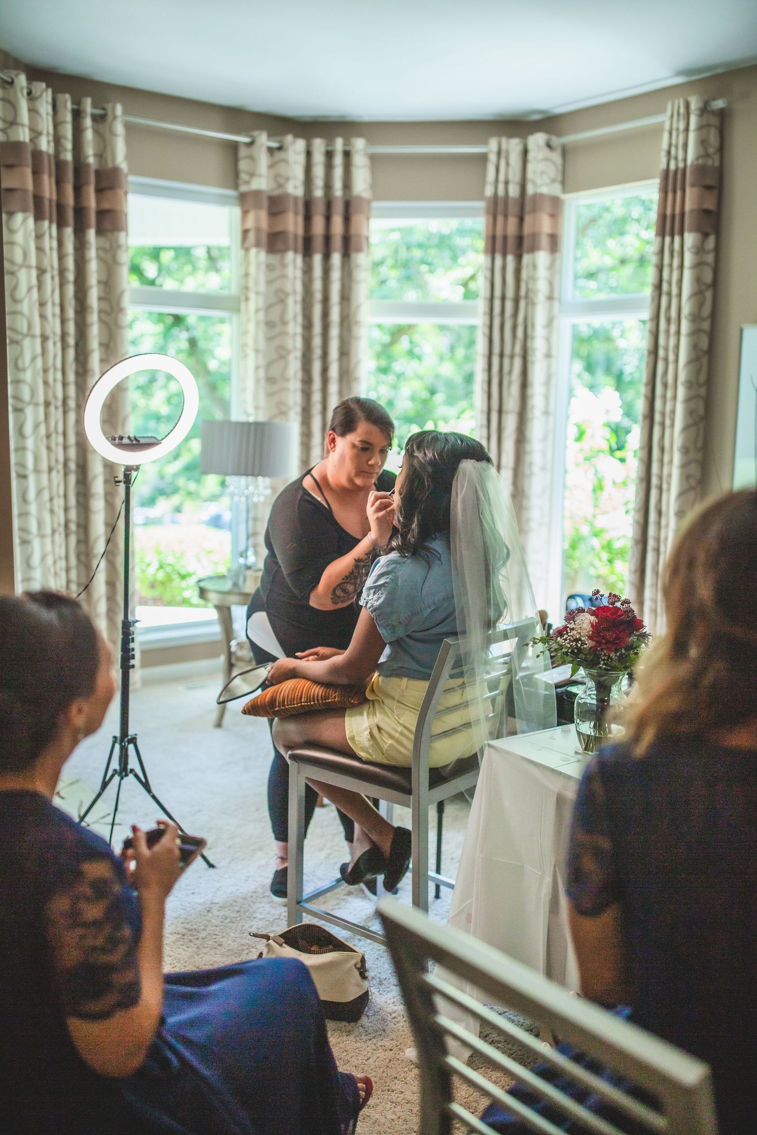 Makeup artist applies cosmetics to someone wearing a veil in a bright room with floral curtains.
