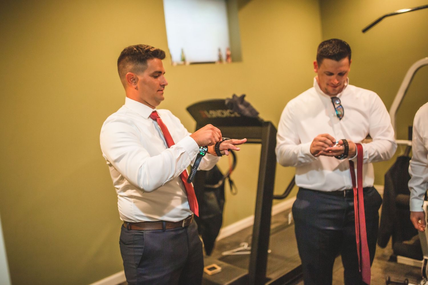 Two people in white dress shirts and red ties prepare for a formal event in a room.