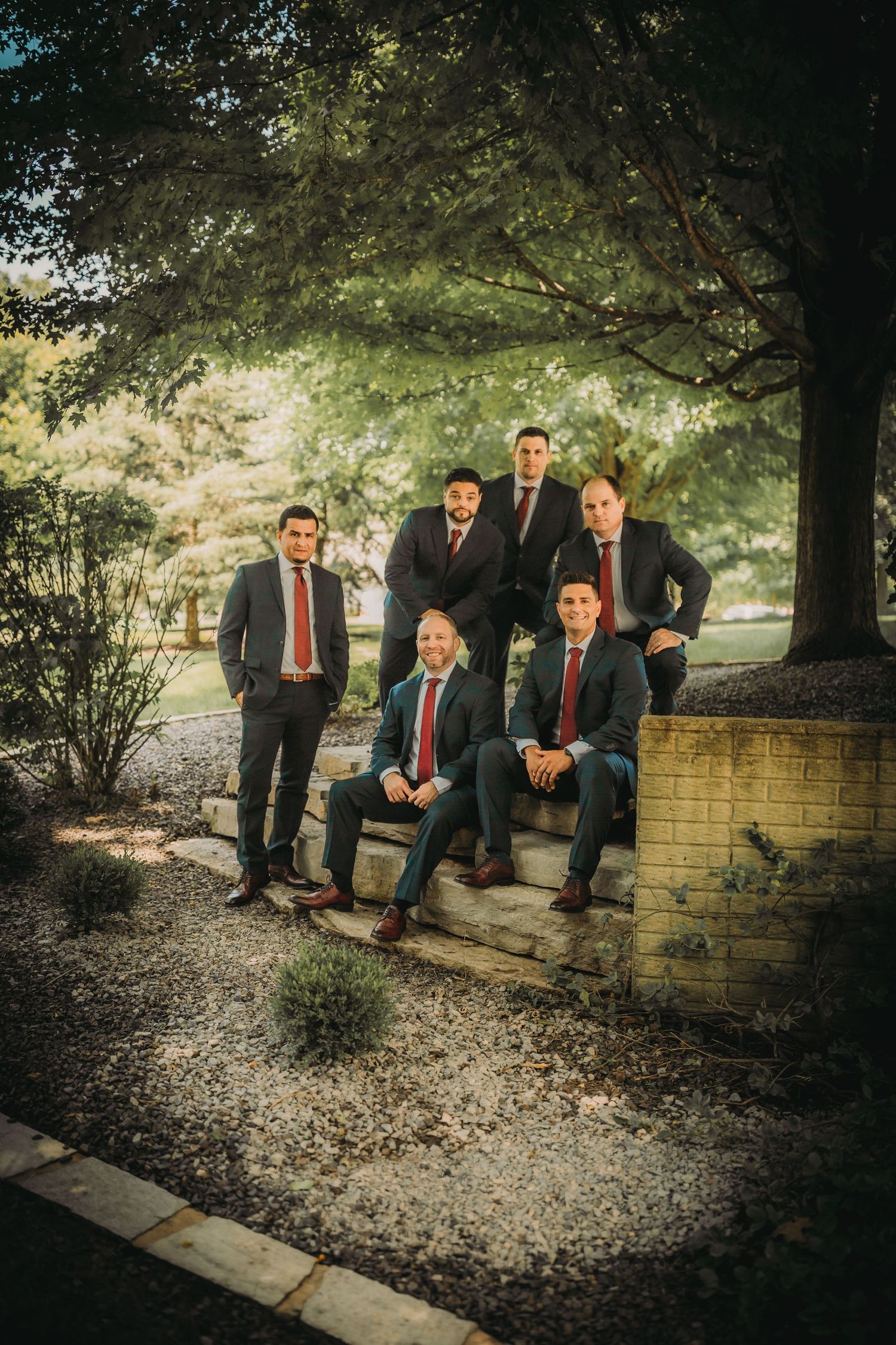 Wedding party in formal attire pose on stone steps under a canopy of trees.
