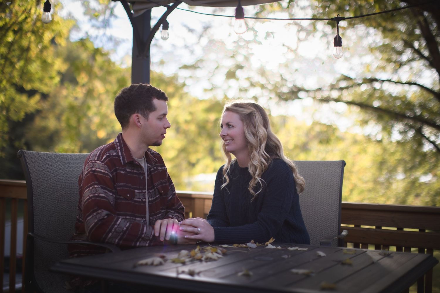 Couple sharing conversation at outdoor table with autumn scenery in background.