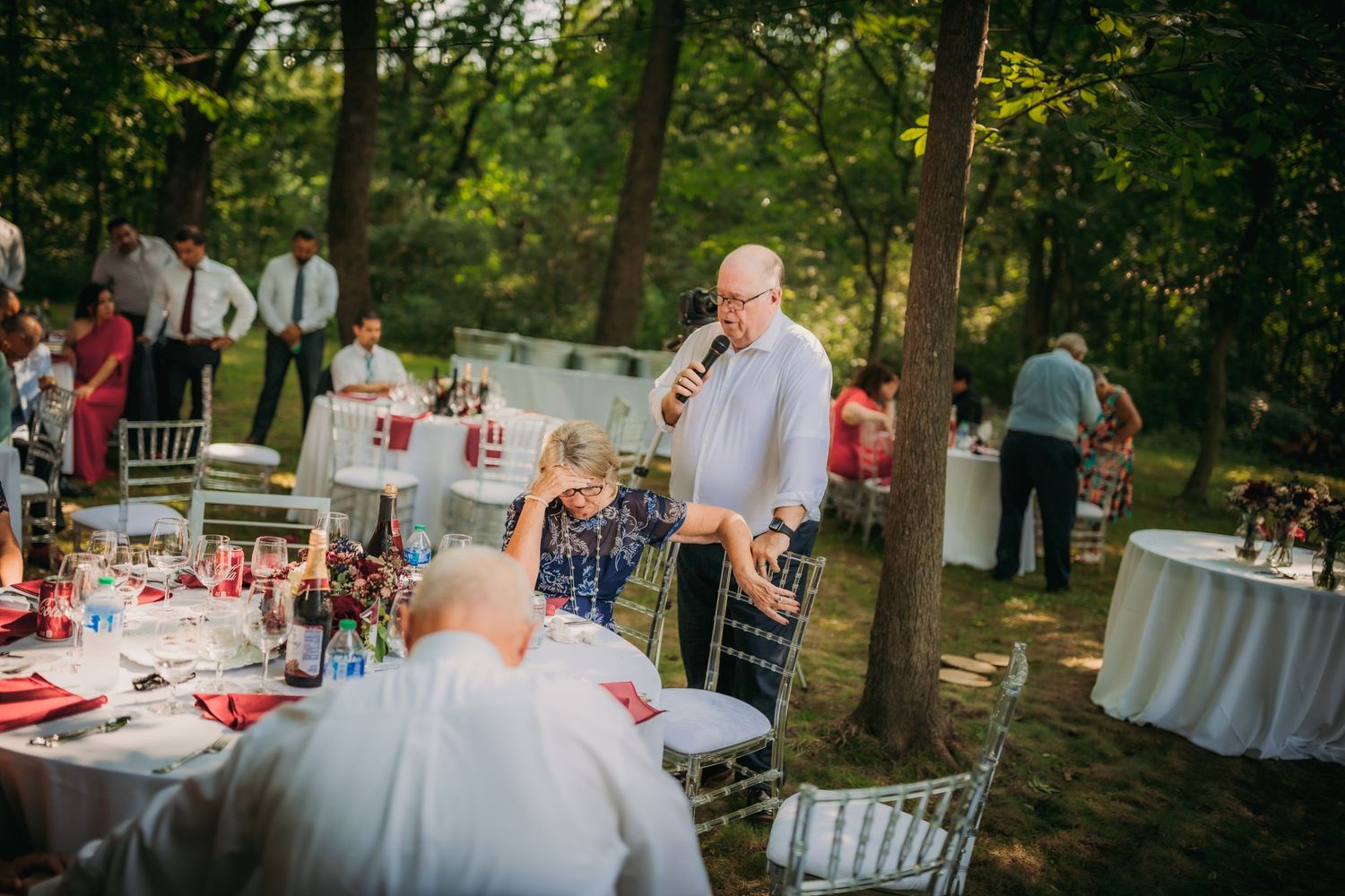 Guests mingle around elegantly set outdoor reception tables under a tree canopy.
