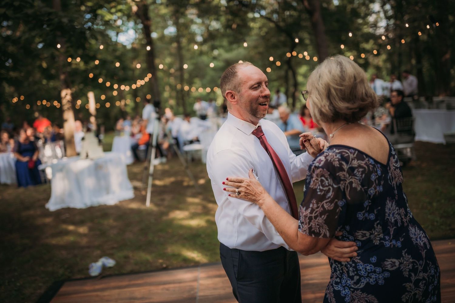 Couples dancing on wooden platform during outdoor reception with string lights and forest scenery.