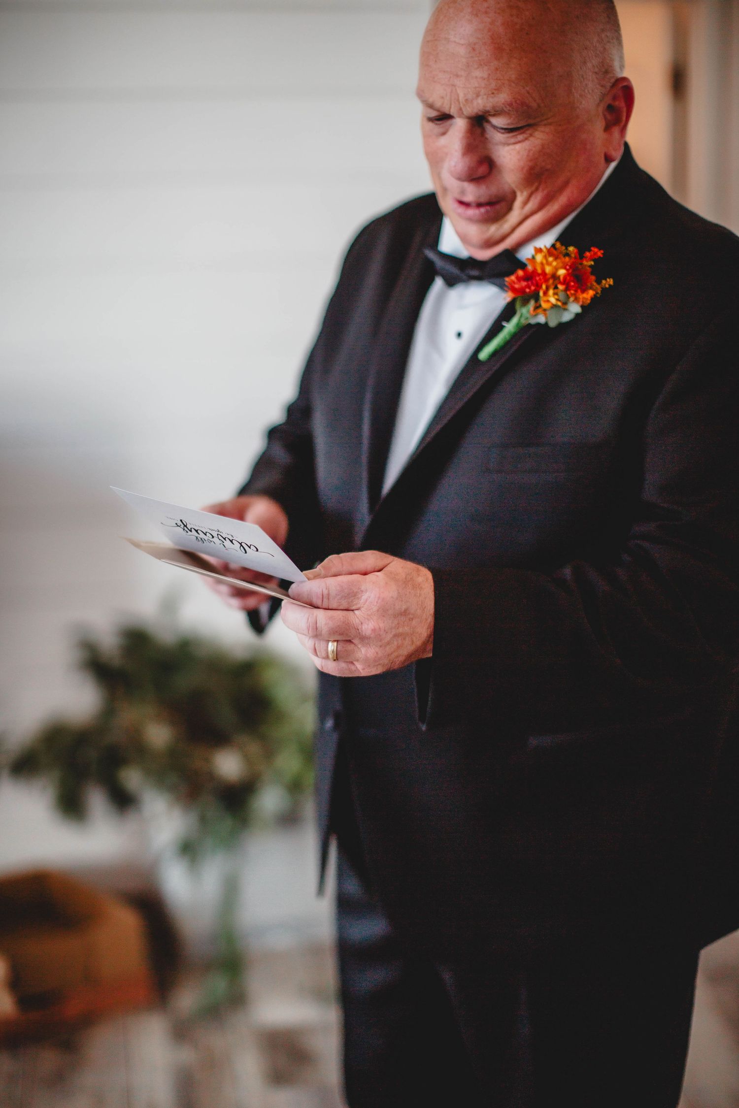 A person in a tuxedo with an orange boutonniere reads a message from a paper at a wedding ceremony.
