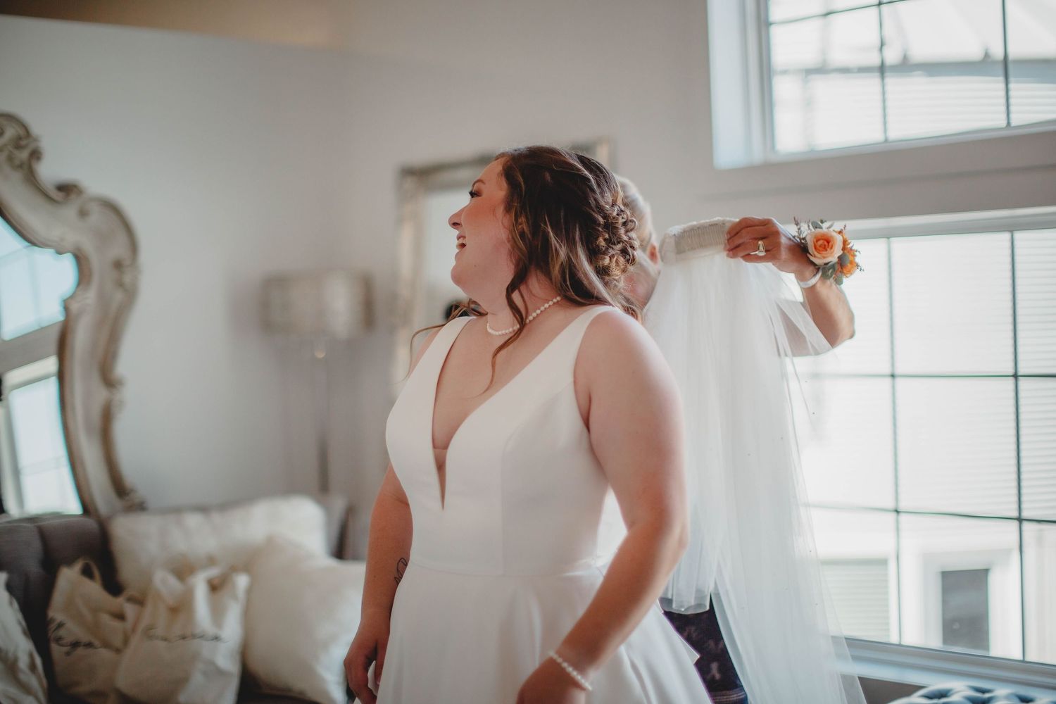 A bride in an elegant white sleeveless gown stands by a window during wedding preparations.