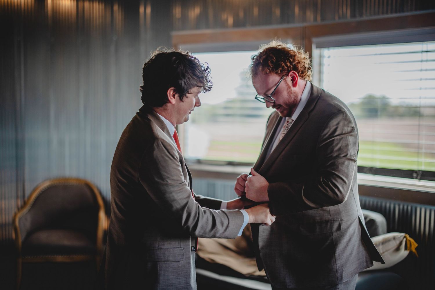 Two people in suits sharing a moment while getting ready in a sunlit room.
