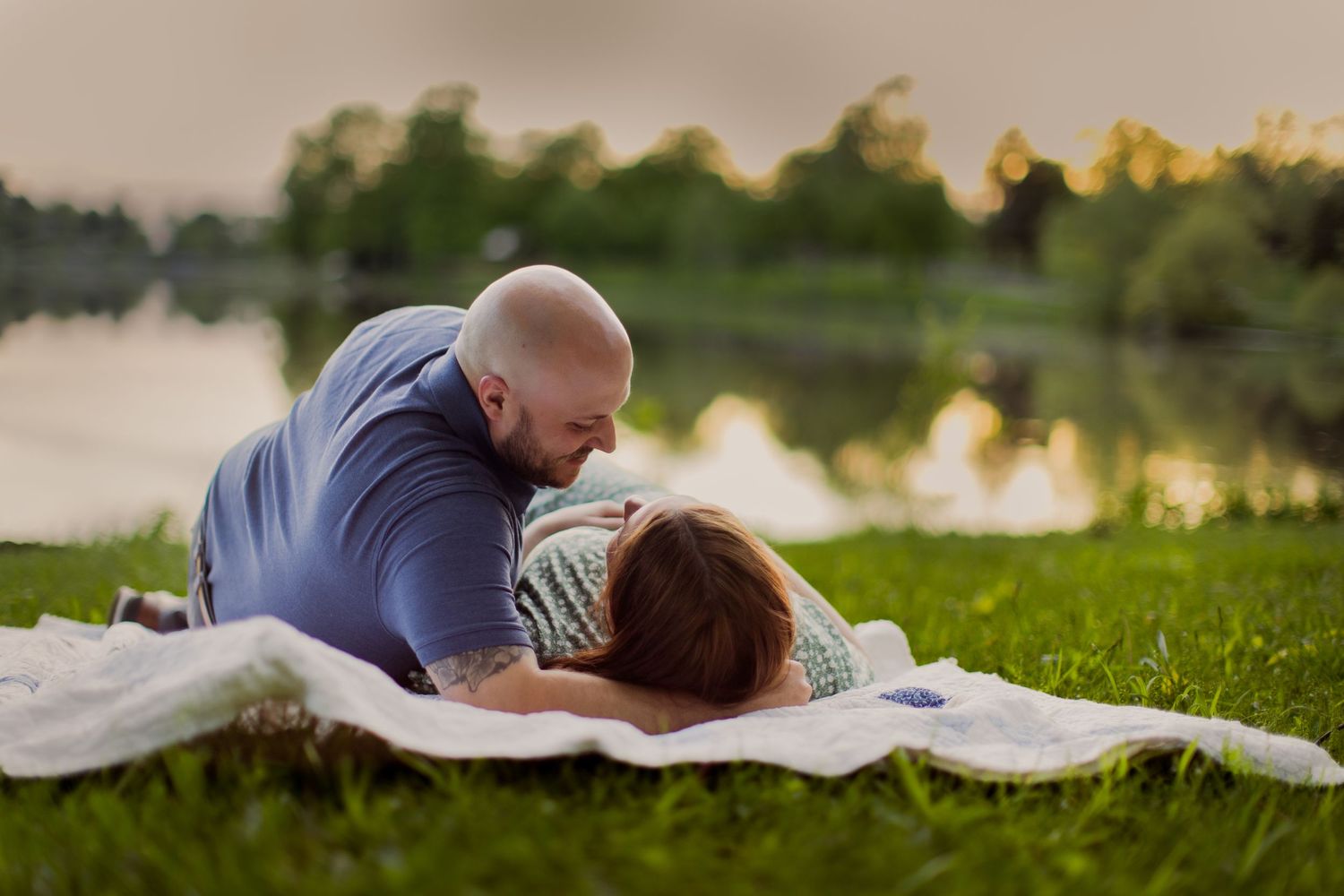 A romantic scene of two people resting on a blanket near a lake at sunset.
