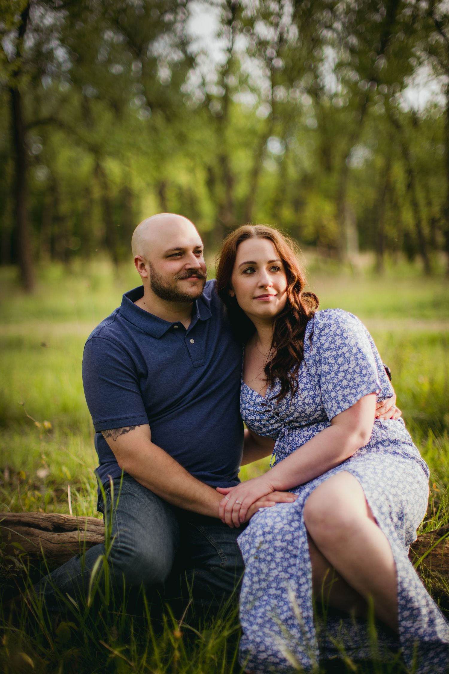 A romantic couple sits together on a fallen log in a green forest setting during an engagement photoshoot.