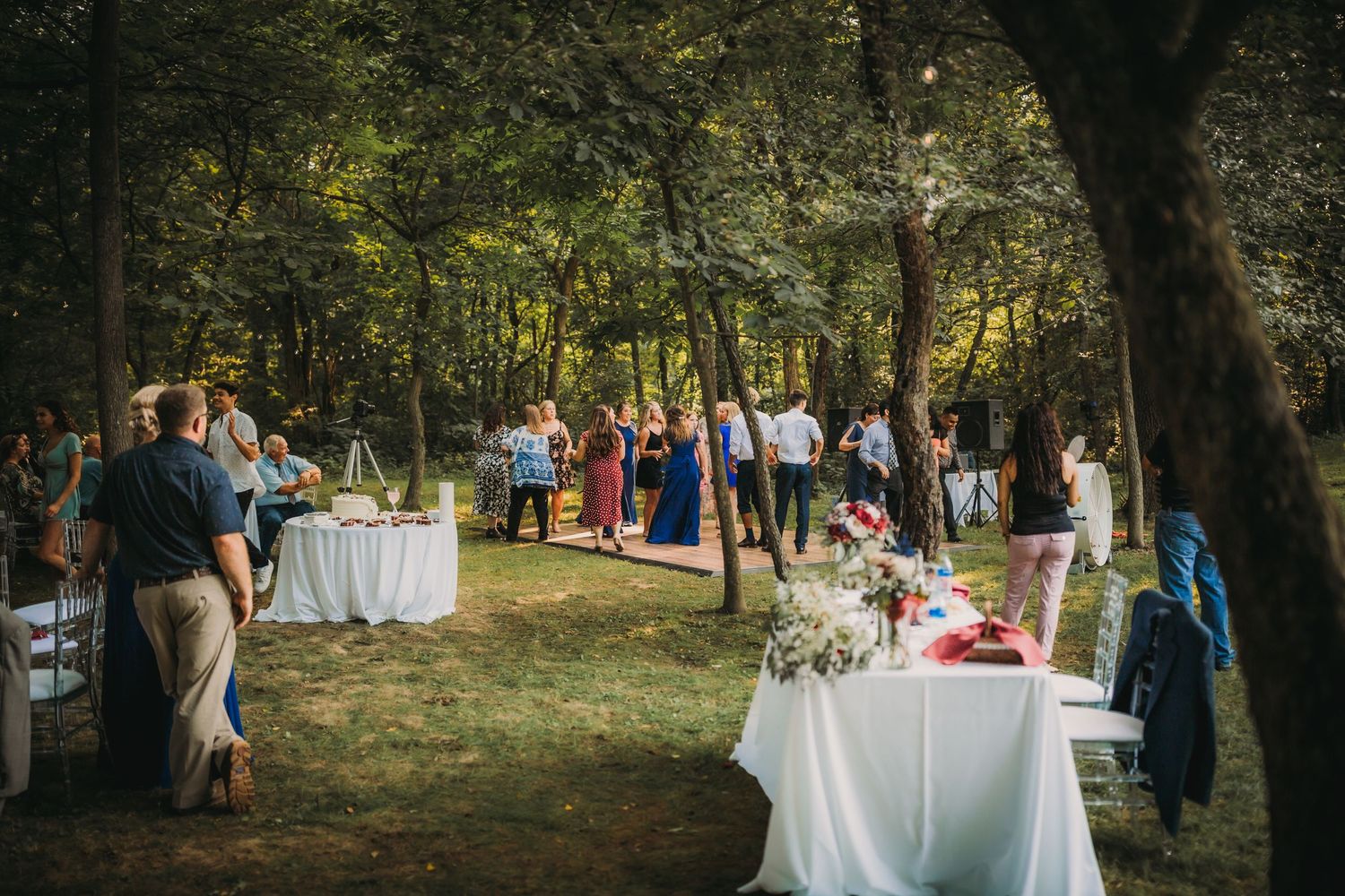 Outdoor wedding reception setup with white tables and guests mingling under tall trees.