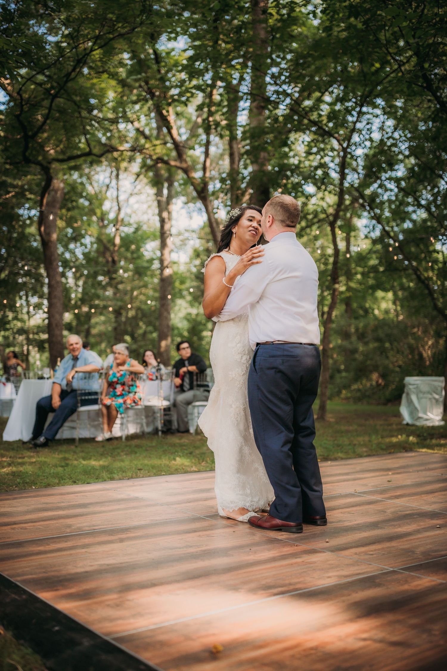 A romantic first dance takes place on an outdoor wooden dance floor with guests seated in the background at a forest wedding.