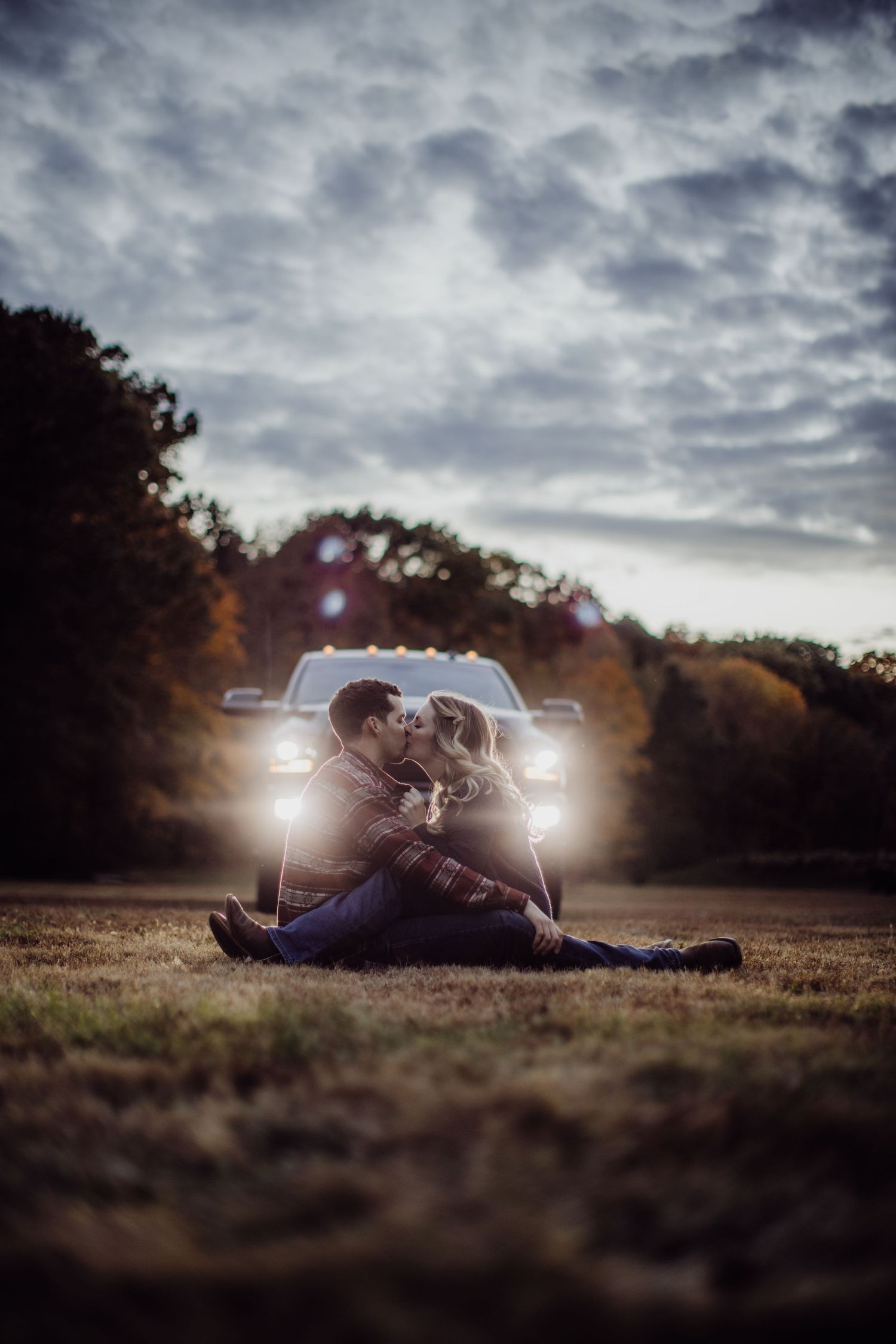 A couple embraces on the ground with dramatic car lighting and moody clouds in the background.