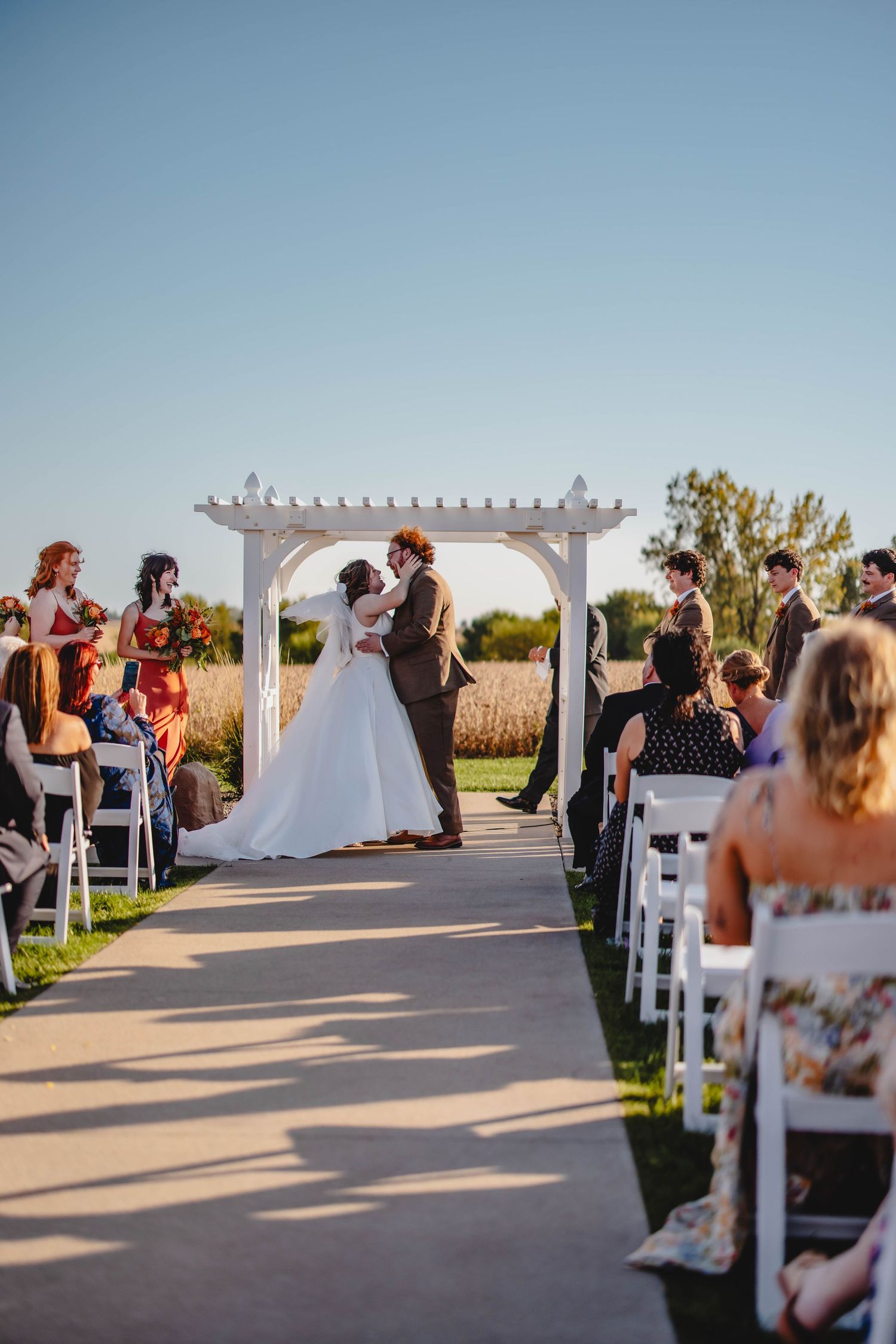 Wedding ceremony at golden hour with white pergola and guests on white chairs.