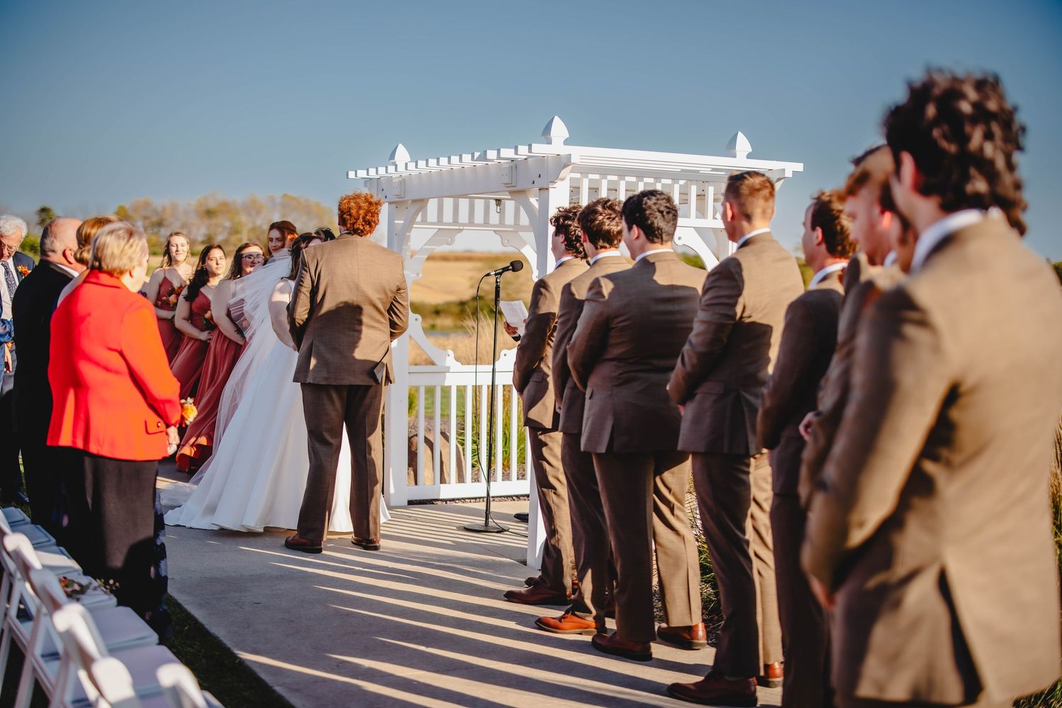 Outdoor wedding ceremony under a white pergola with guests gathered on a sunny day.
