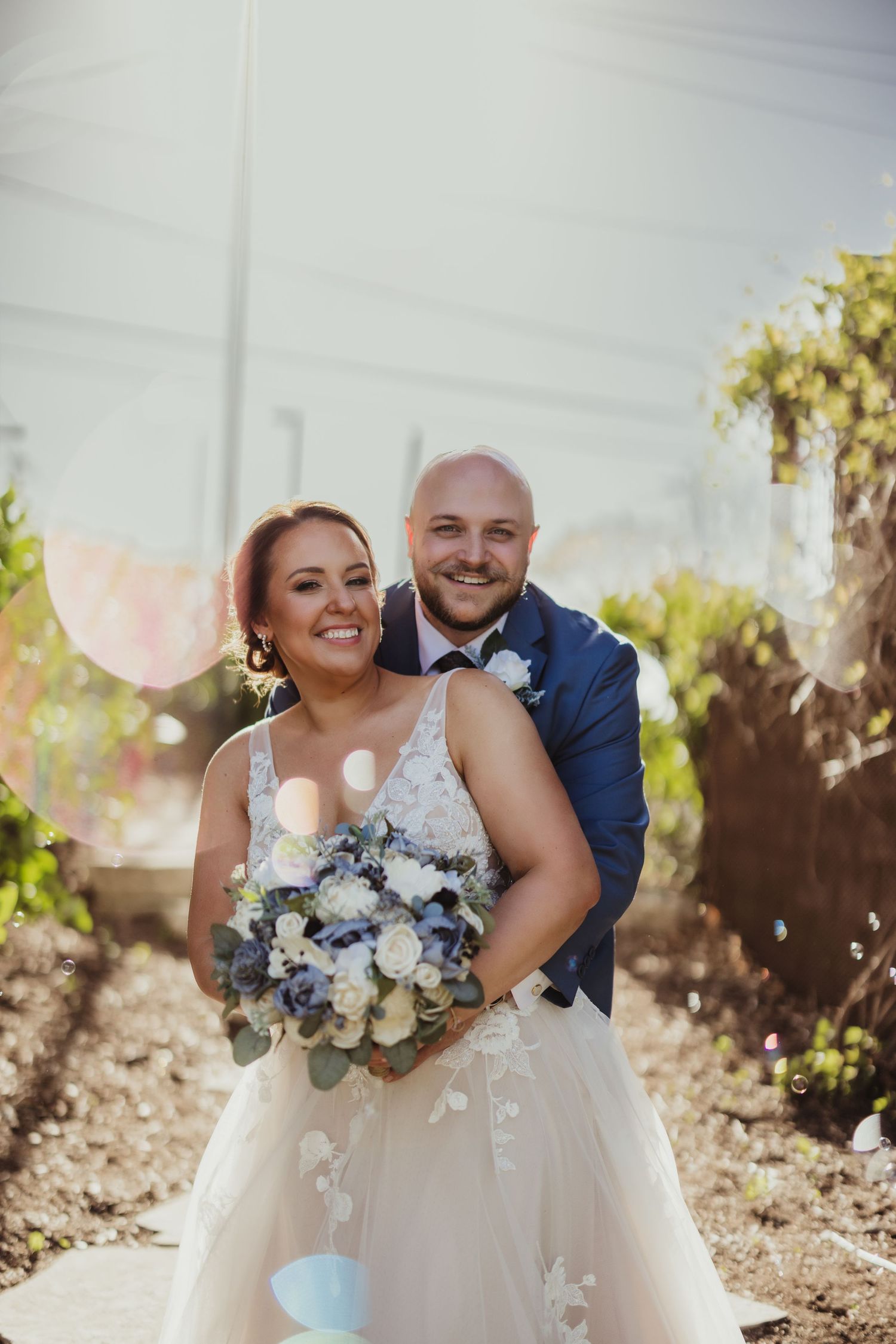 A couple in wedding attire poses together outdoors with a bouquet of blue and white flowers.