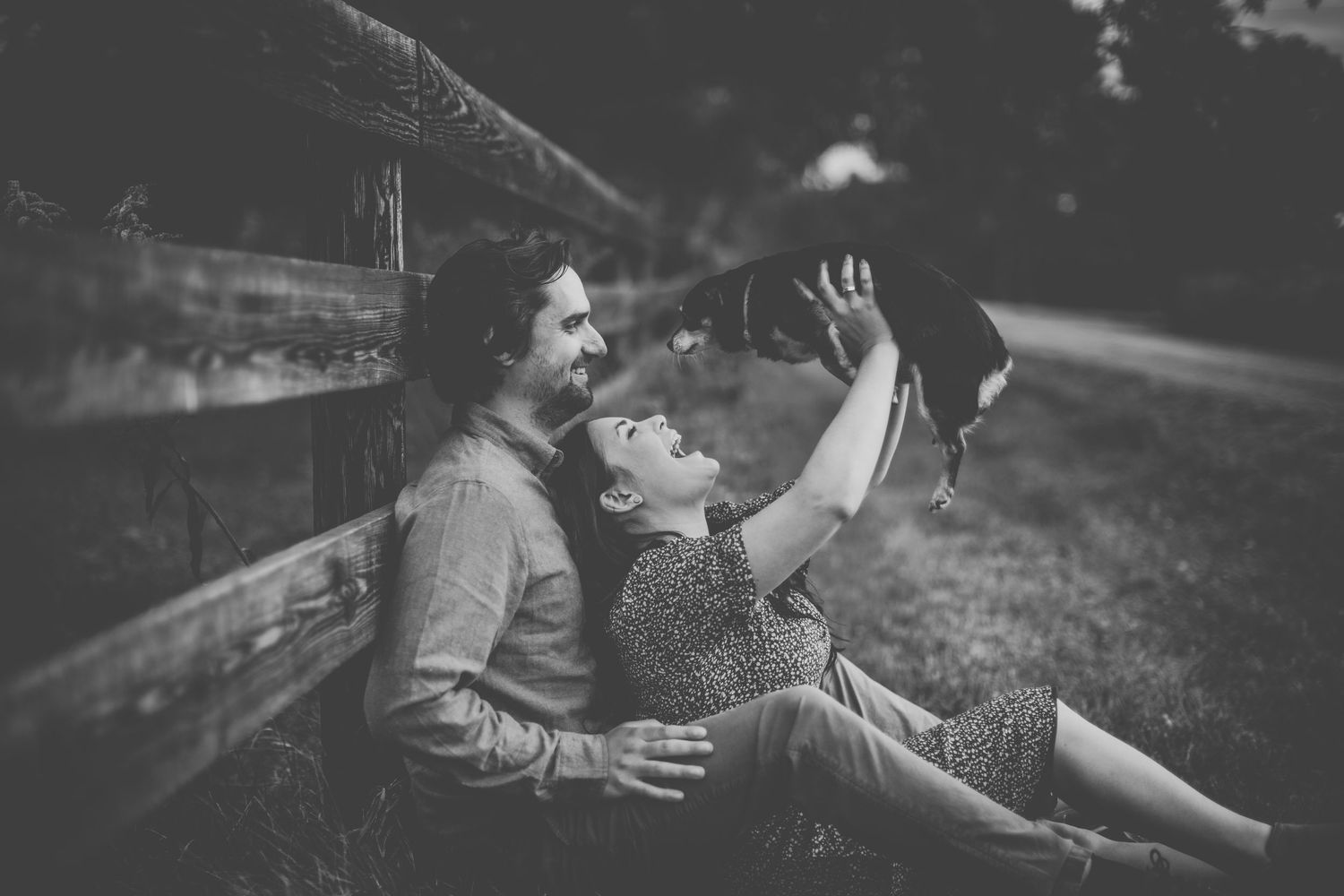 Black and white photo of couple playing with cat while sitting against wooden fence.
