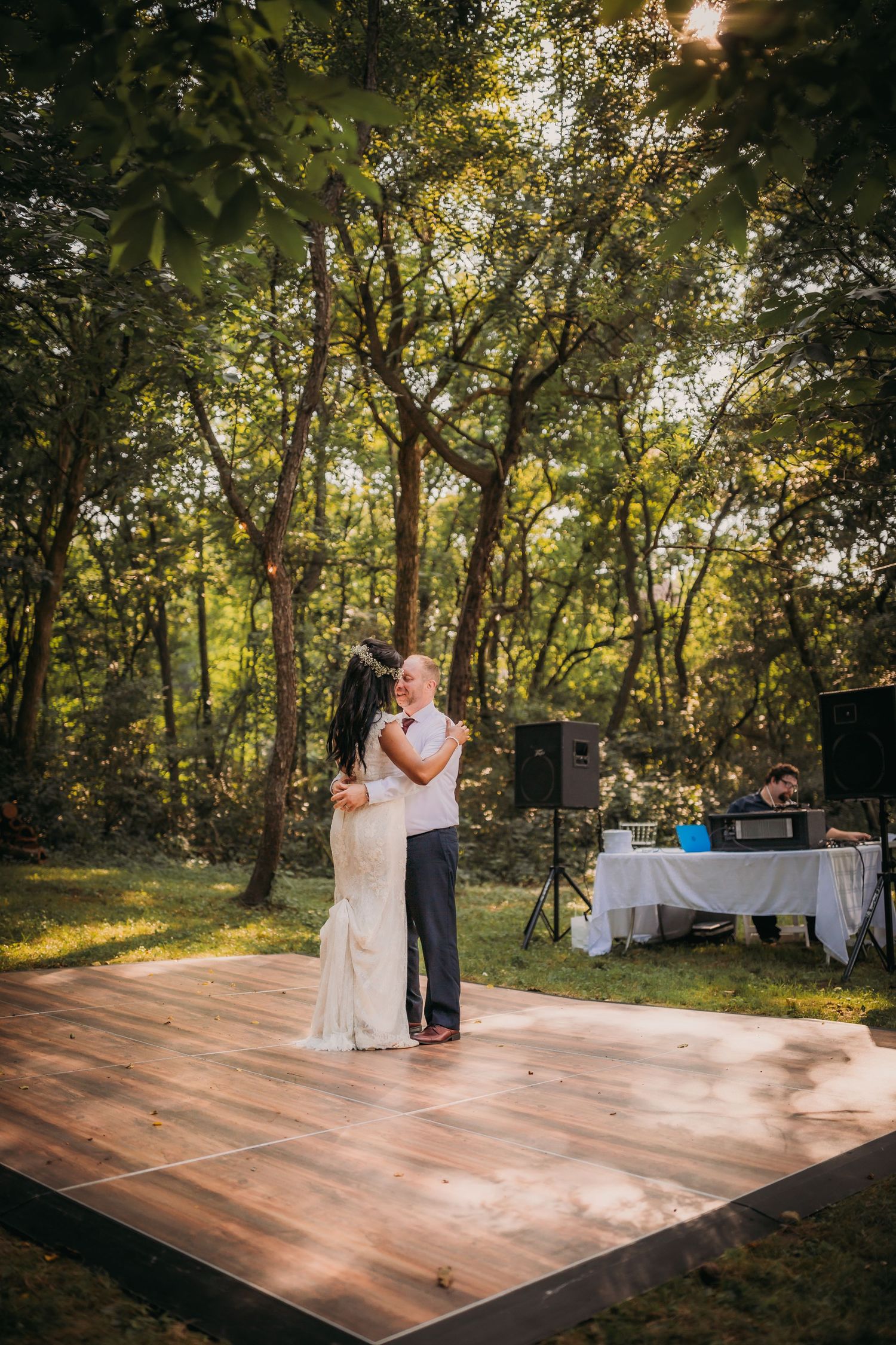 A couple shares their first dance on a wooden platform in a sunlit forest setting.