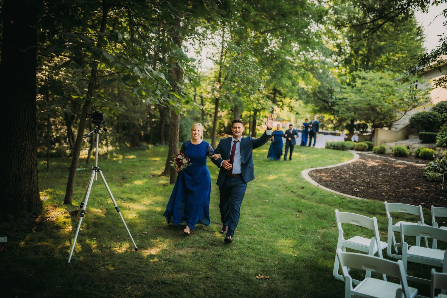 Attendants in royal blue and navy walk along a landscaped garden path at an outdoor ceremony.