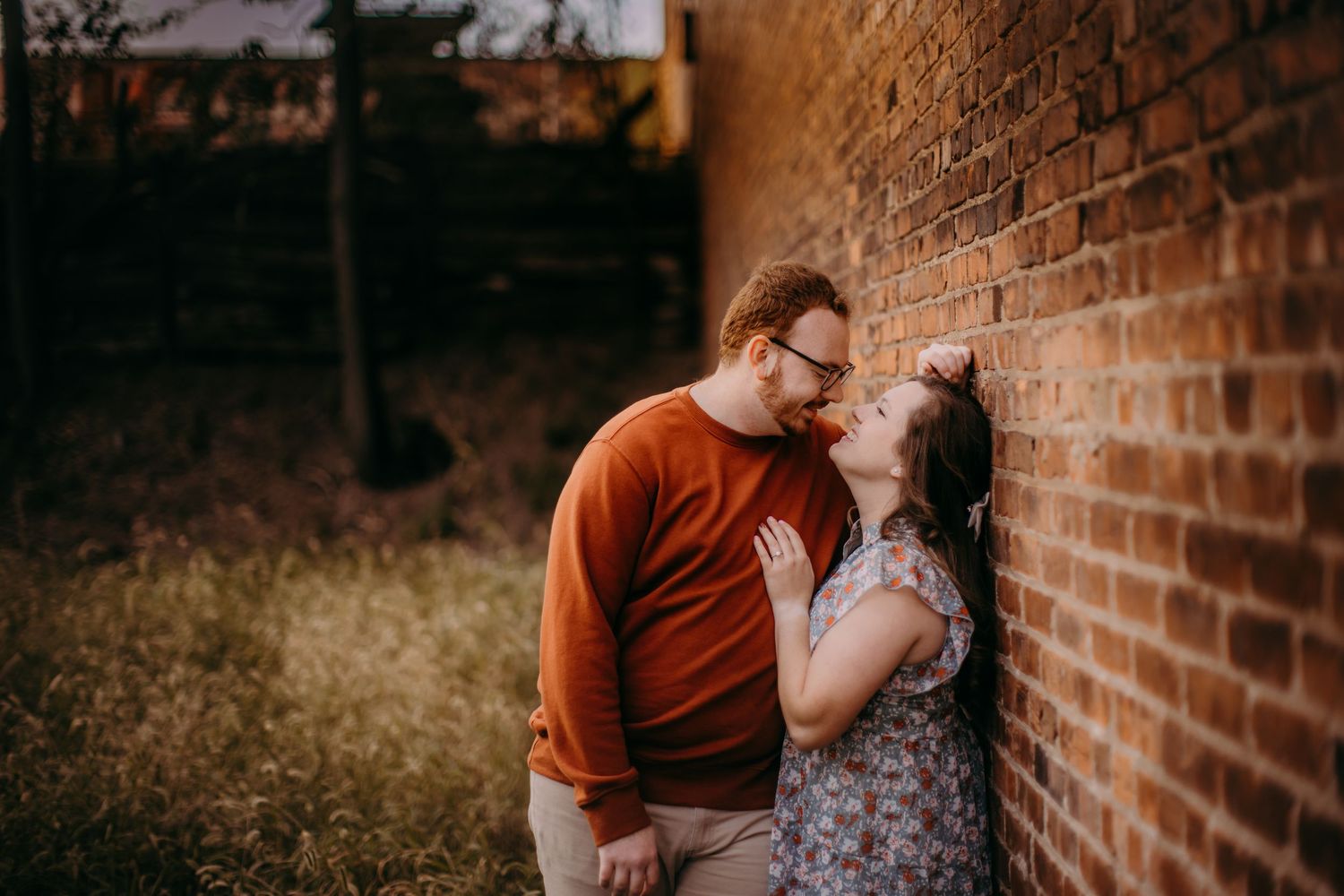 An intimate engagement photo session features a couple embracing by a textured brick wall.