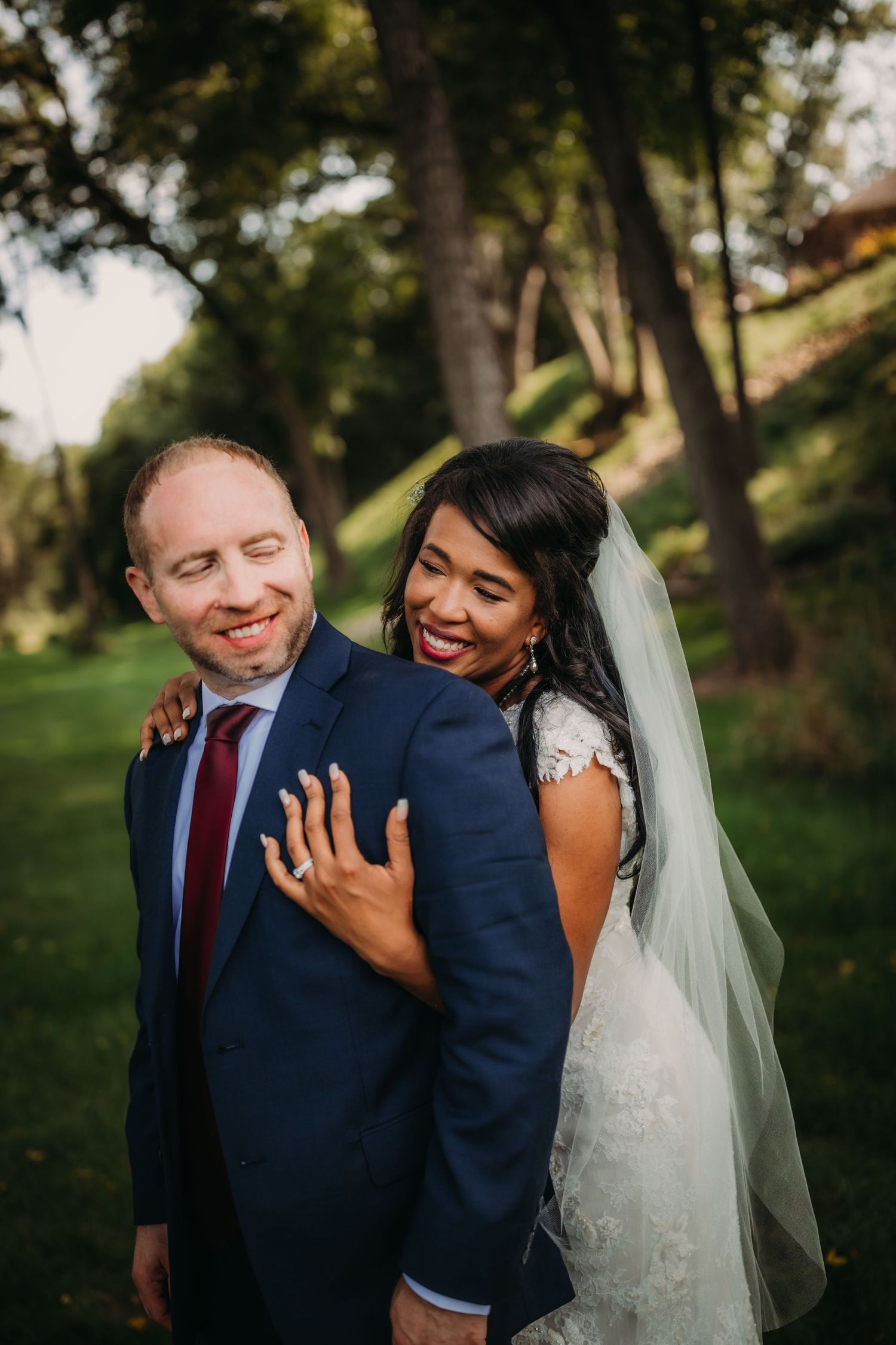 A bride in white lace dress and groom in navy suit share a playful moment outdoors.