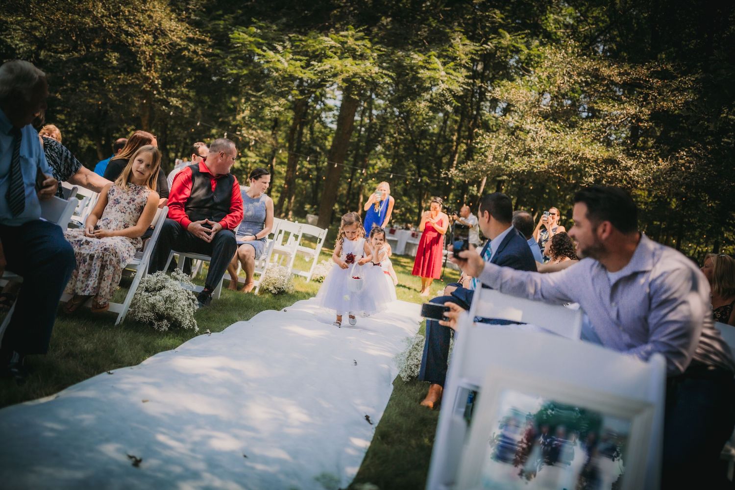 A small child in white walks down a white aisle runner at an outdoor wedding ceremony.