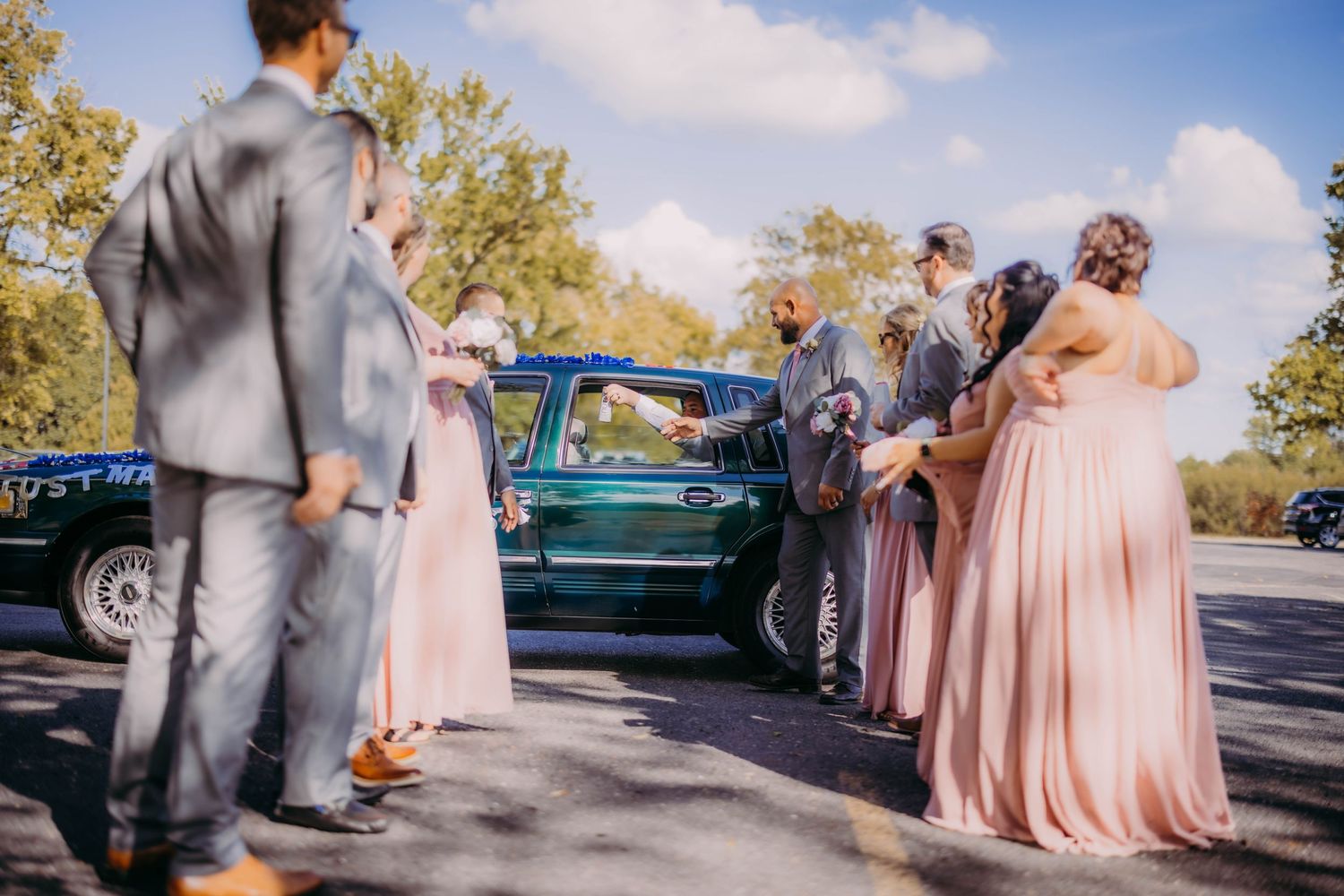 Wedding party in pink and grey attire gathered in parking lot on sunny day.