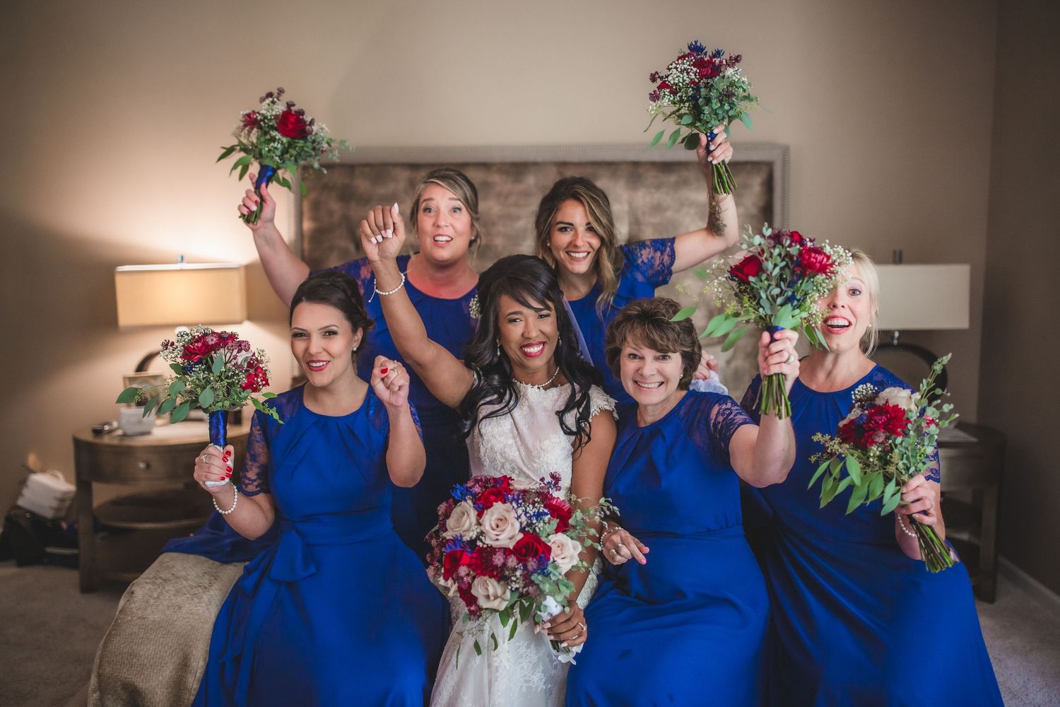 Bridal party in blue dresses raise floral bouquets while posing in bedroom.