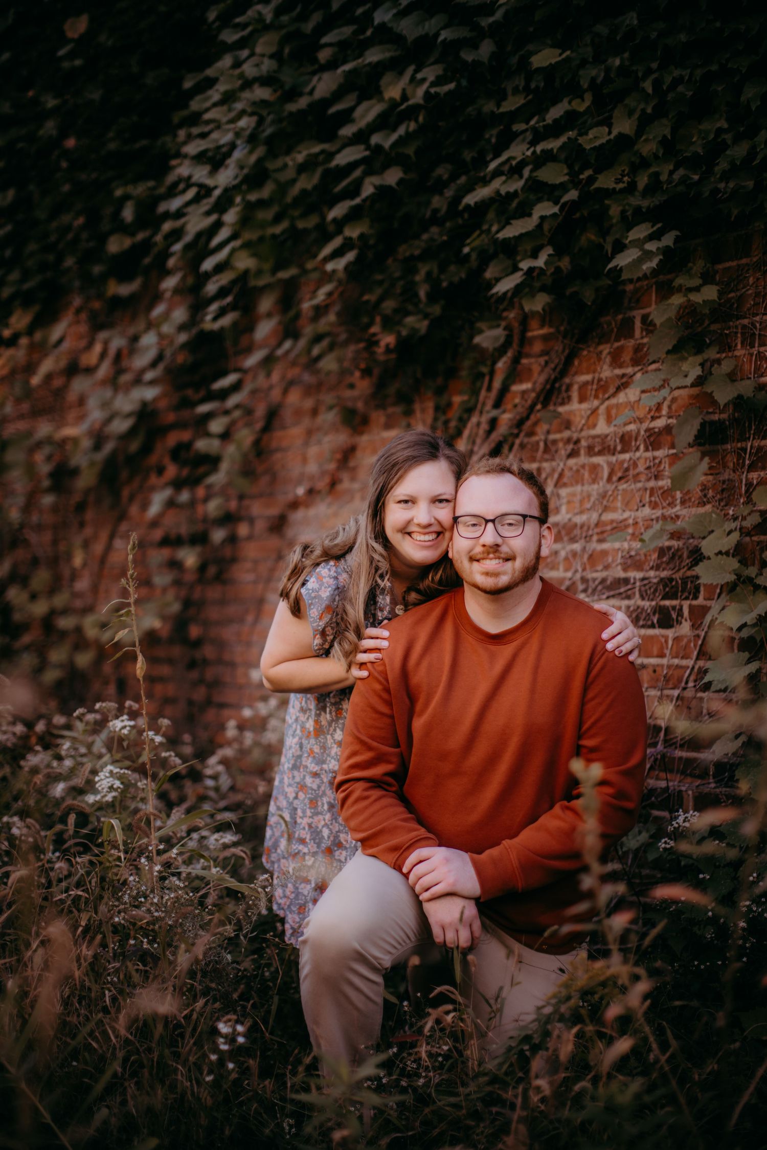 Two people pose playfully in front of a weathered brick wall during a casual outdoor photo session.