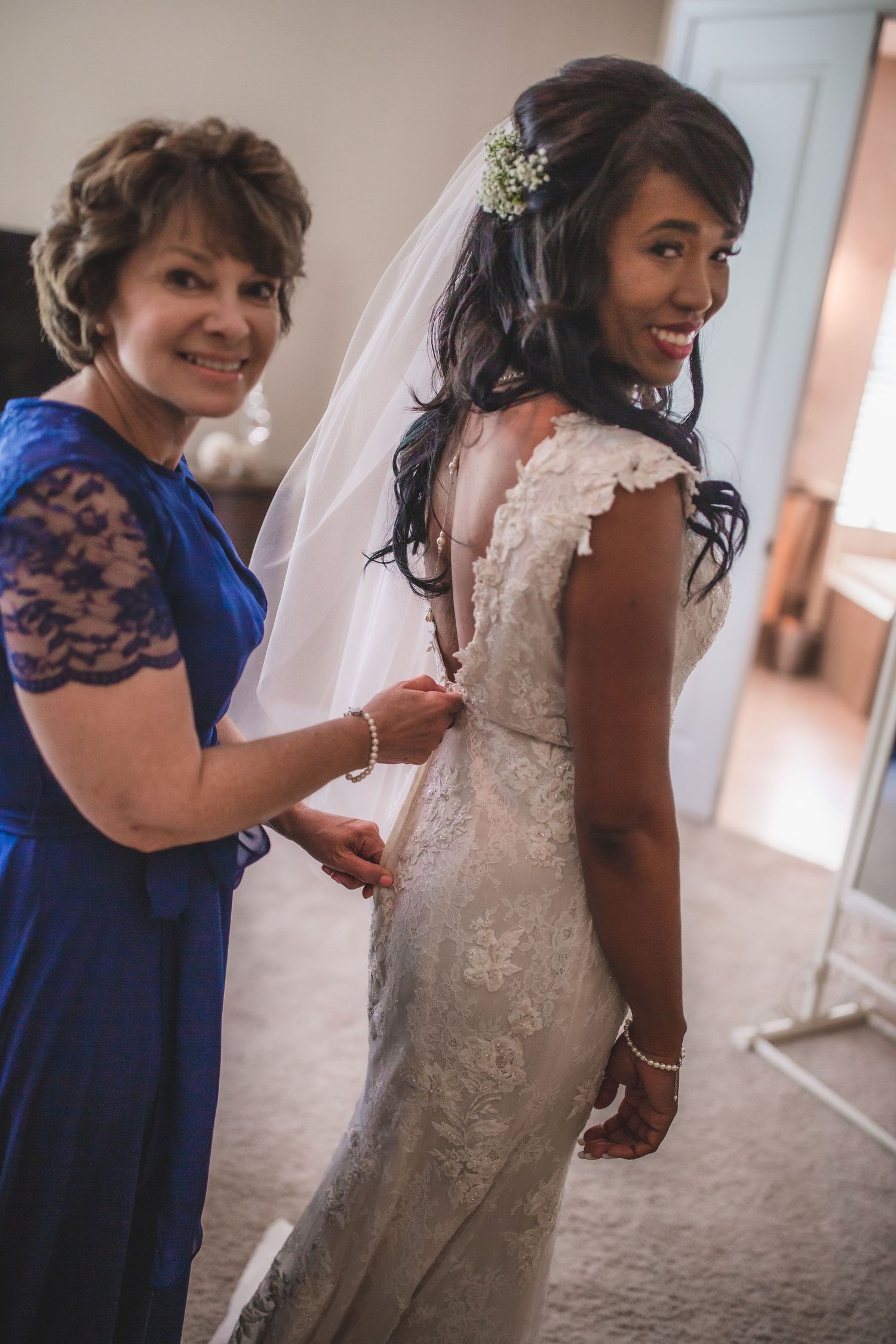 A bride in a lace wedding dress gets assistance with her gown from a woman in a blue lace dress.