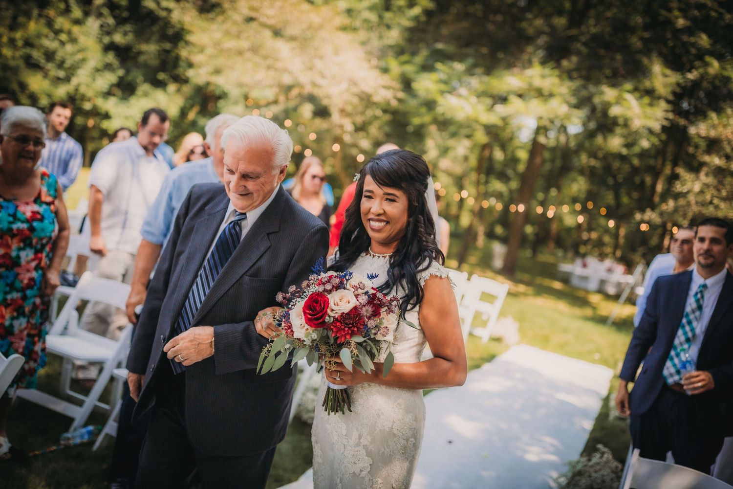 Guests celebrate during an outdoor garden wedding ceremony under towering trees with white folding chairs.