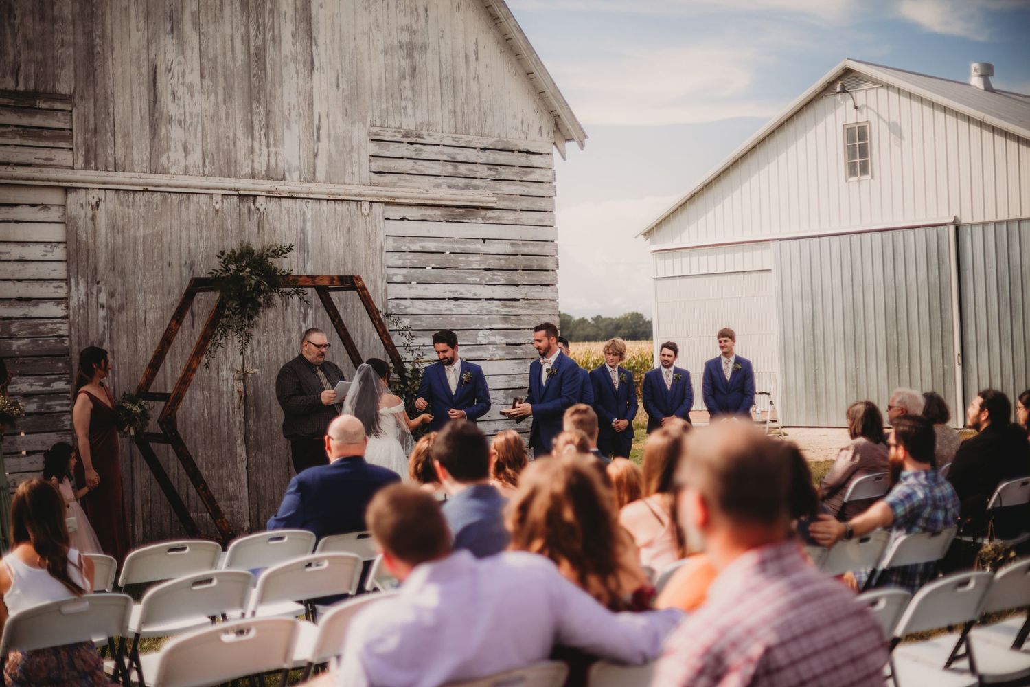 Outdoor wedding guests seated in front of weathered white barn for ceremony.