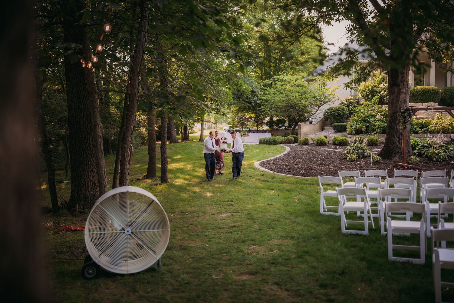 White ceremony chairs arranged in rows on a green lawn surrounded by mature trees.