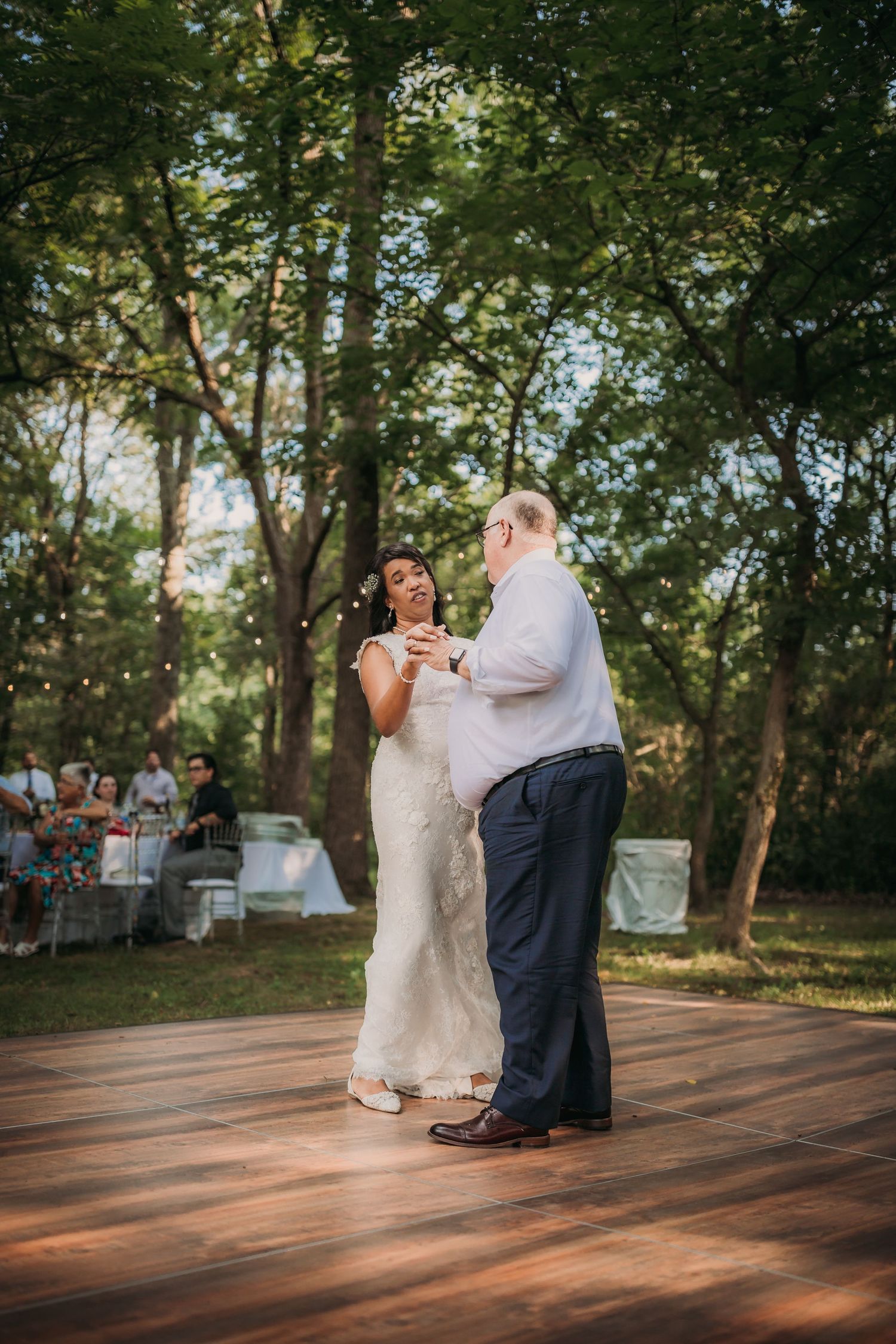 Two people share a tender moment while dancing on a sunlit wooden dance floor at an outdoor wedding venue.