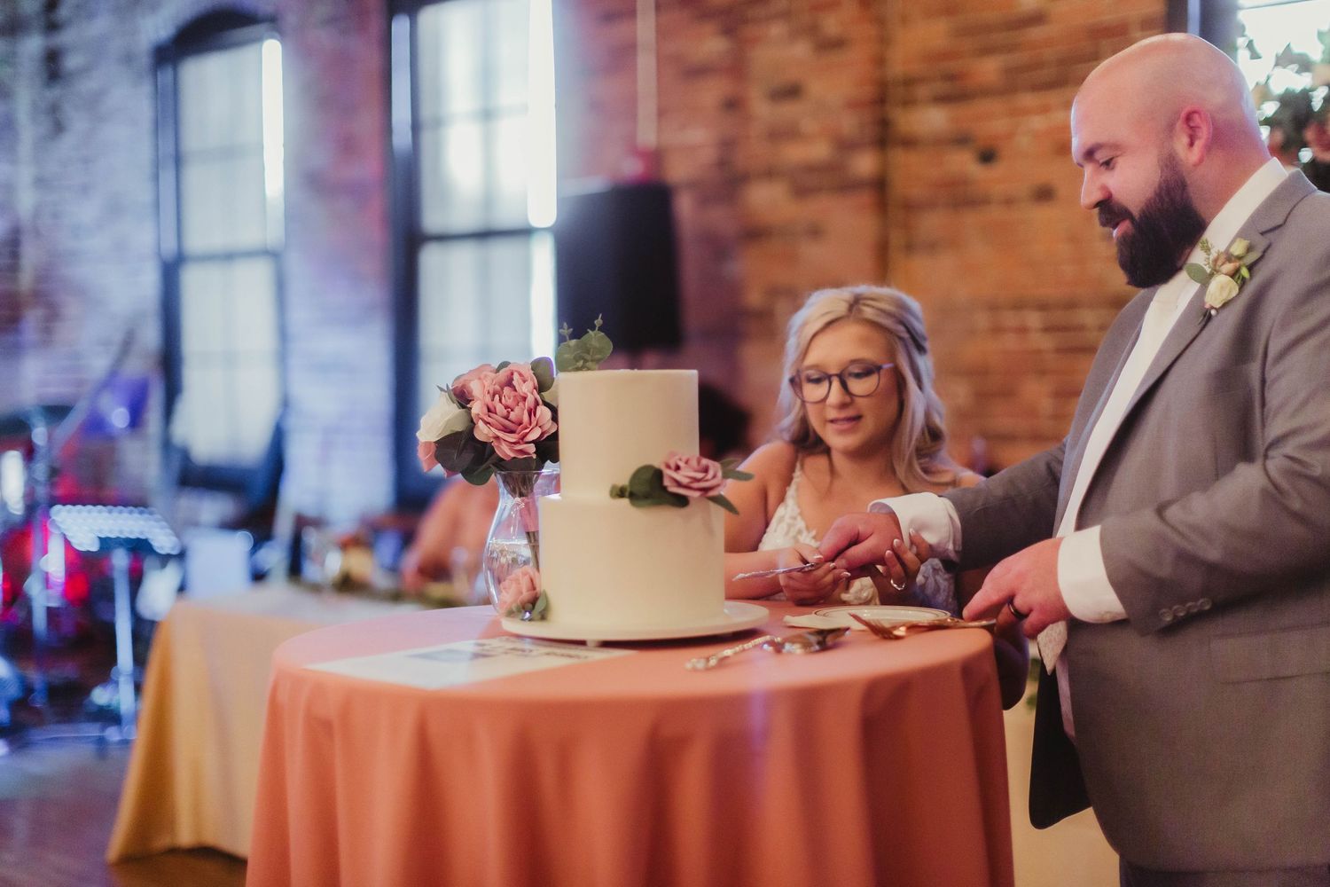Two-tiered white wedding cake being cut on orange tablecloth during reception.