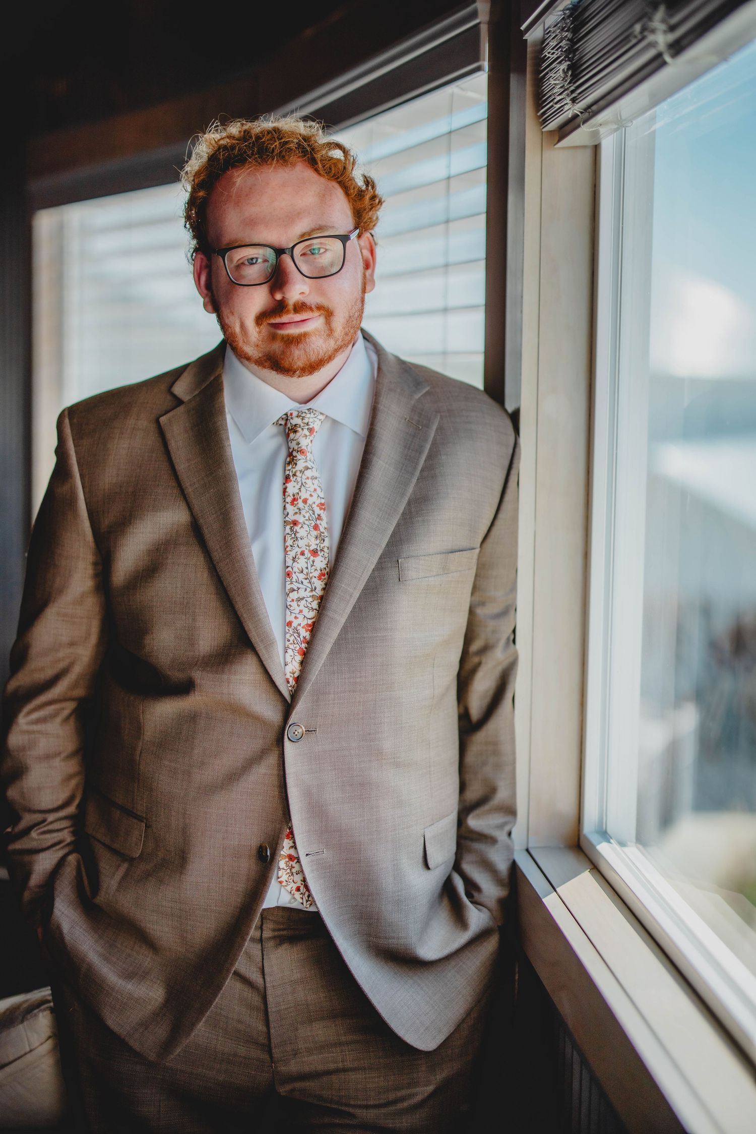 Professional portrait of someone in formal attire standing by a bright window.