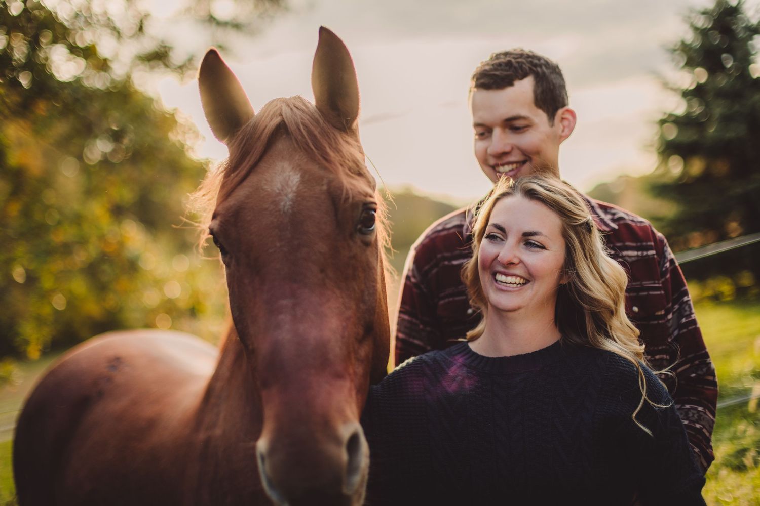 Horse and couple share tender moment in sunlit meadow during sunset.
