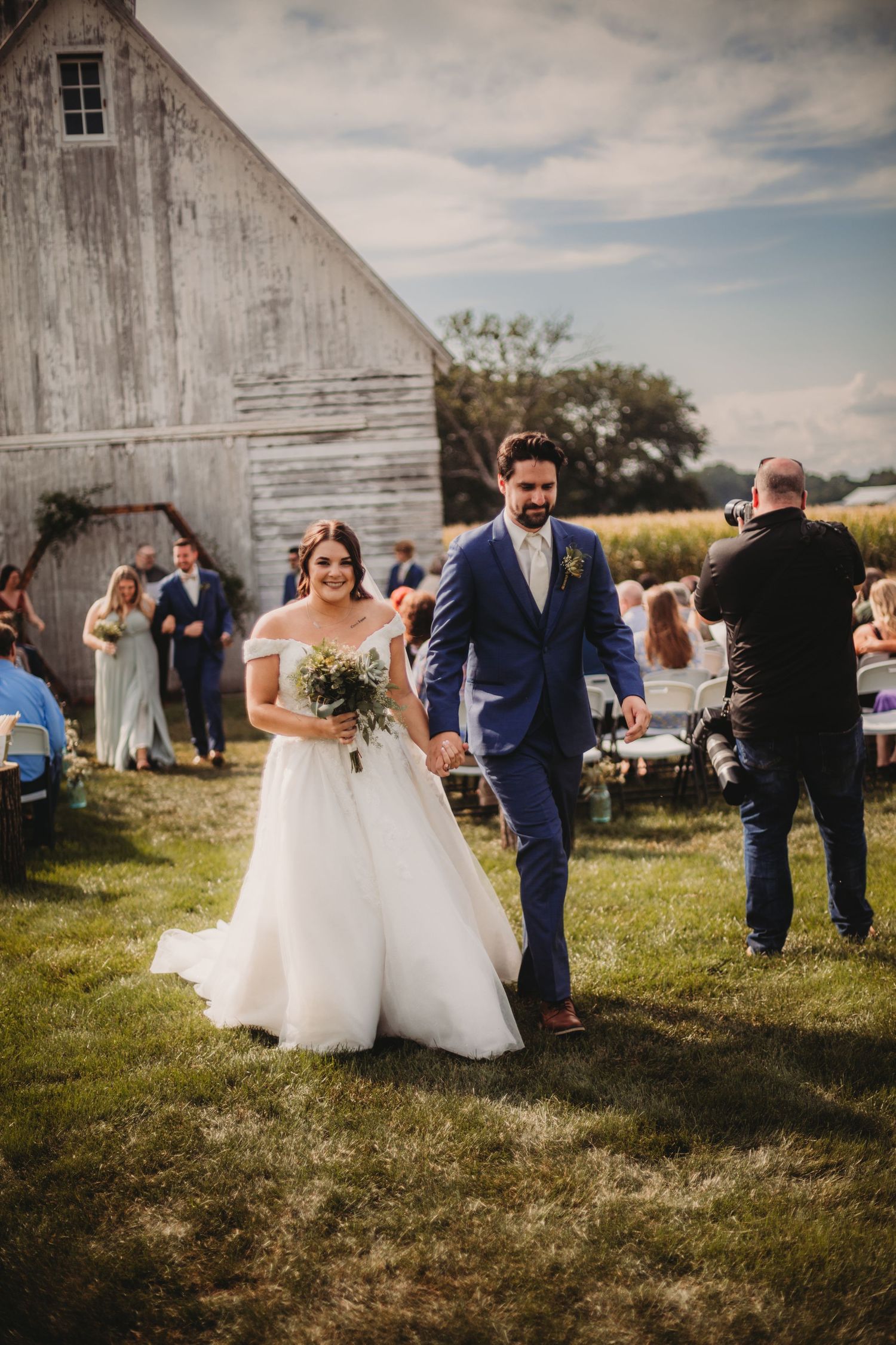 Wedding party poses for photos outside rustic white barn on grassy field.