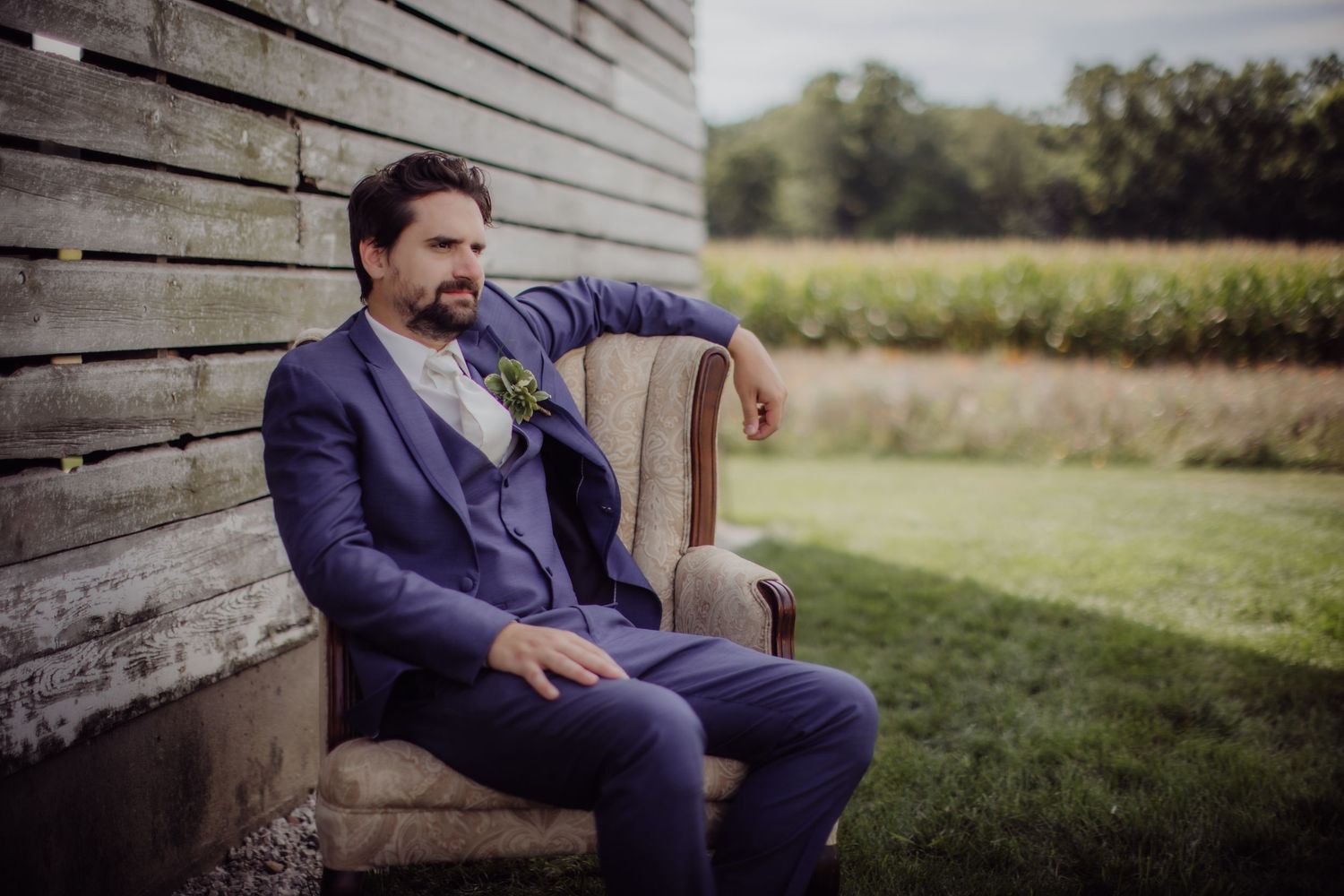 Groom in navy blue suit seated in vintage chair against rustic barn wall.