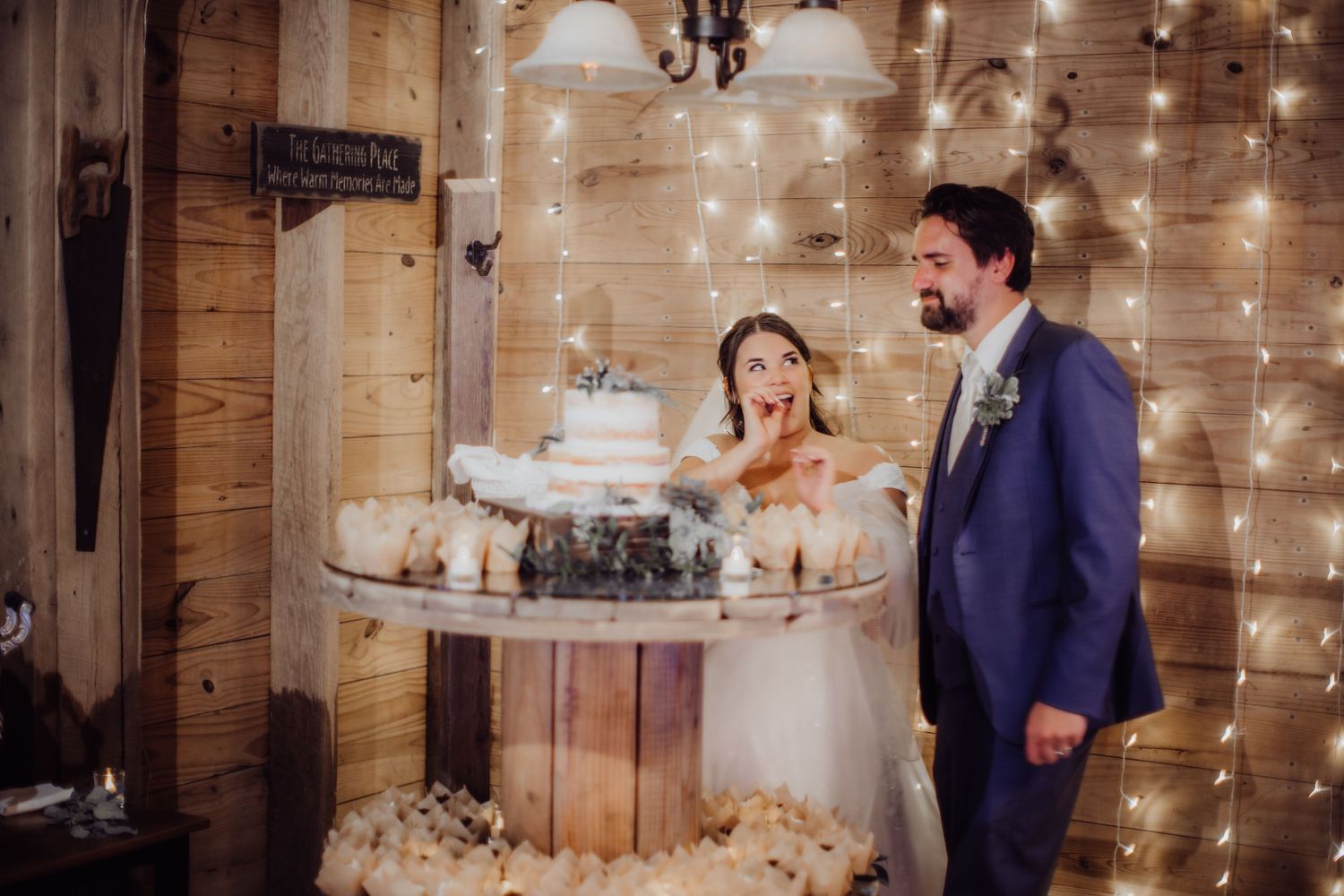 Newlyweds share a playful moment while cutting their wedding cake in a rustic barn venue with twinkling lights.