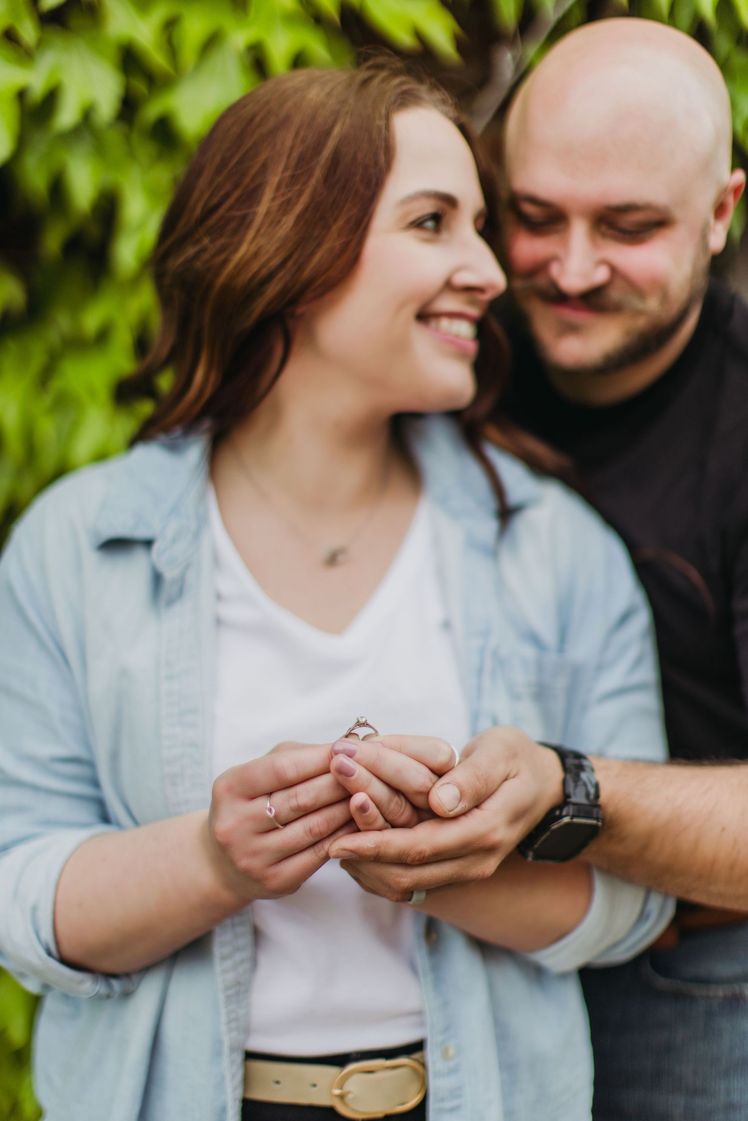 Couple shares intimate moment while examining ring together against green foliage background.