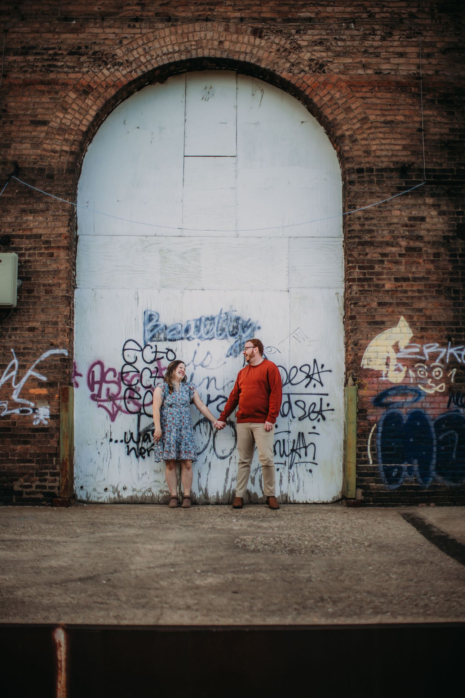 A couple poses together in front of a graffiti-covered archway in an urban setting.