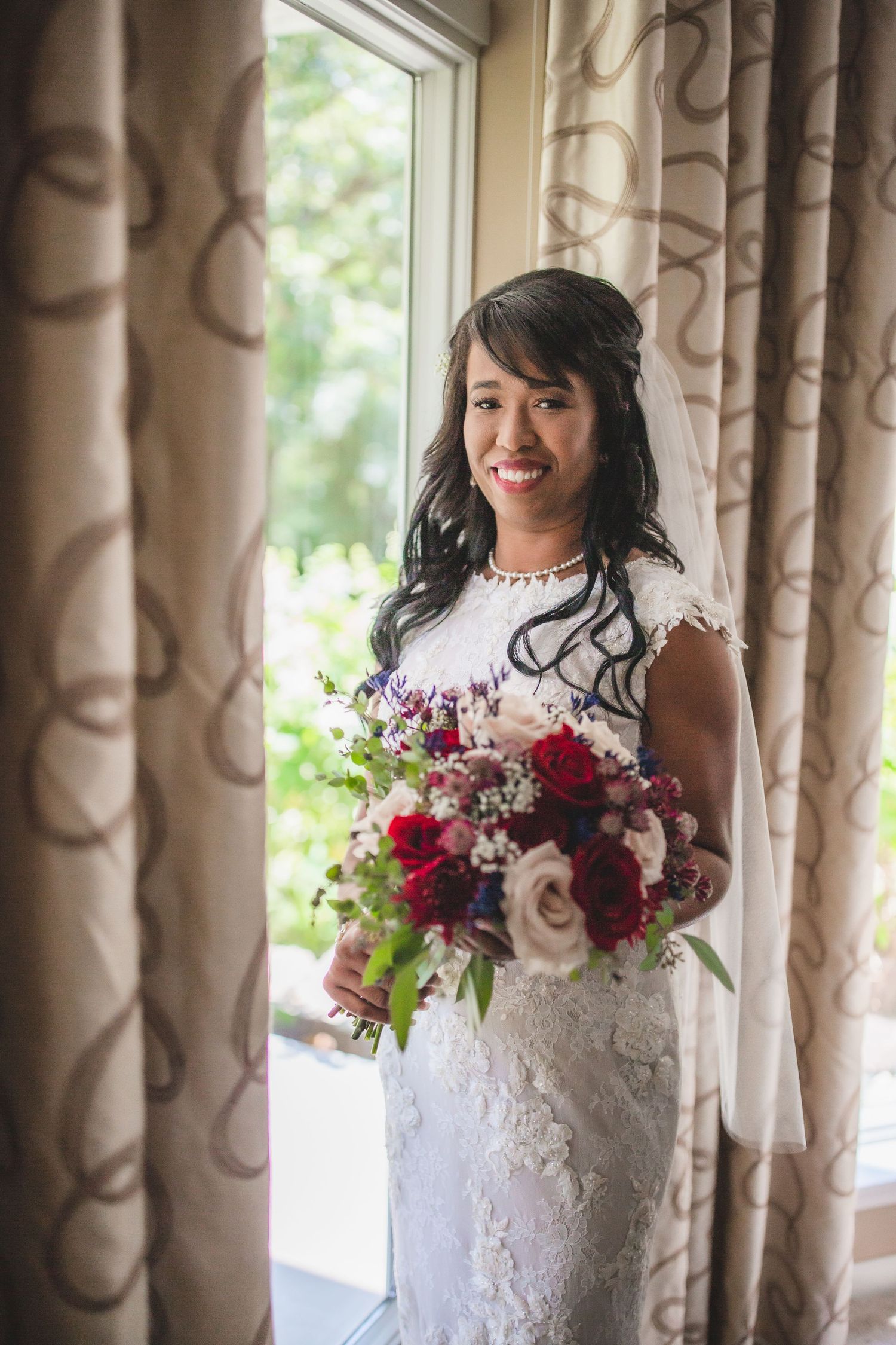 Elegant bride in lace dress holds burgundy bouquet against window with patterned curtains.