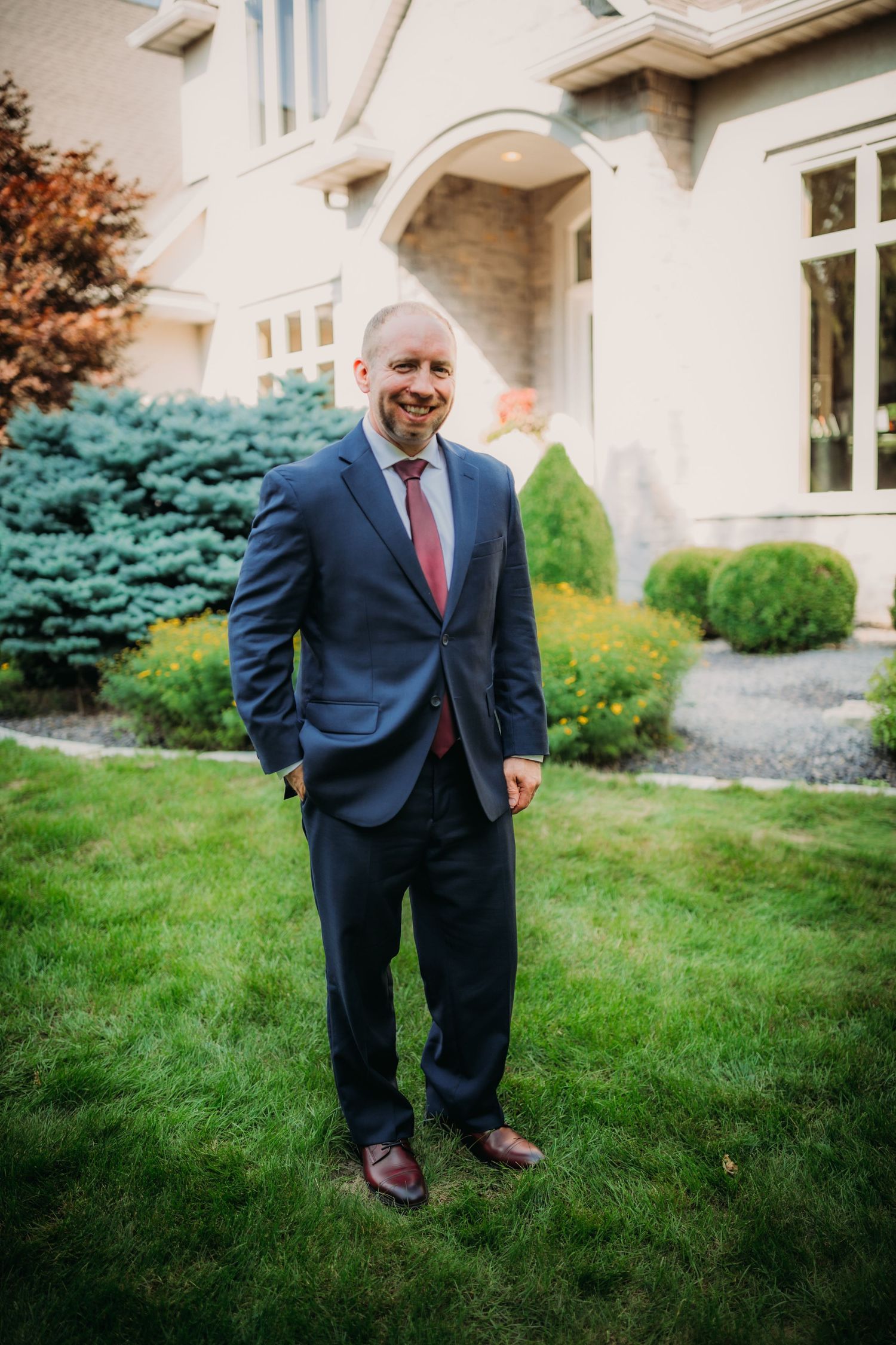 Wedding attire features navy suit with burgundy tie against cream-colored mansion exterior.