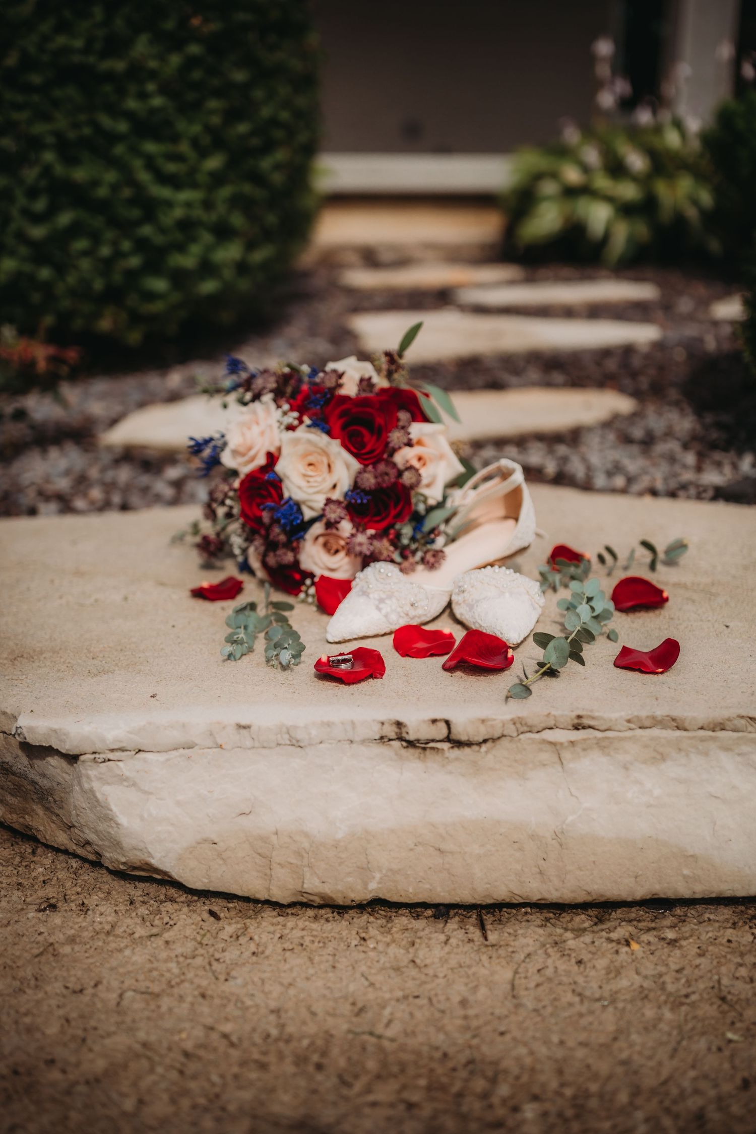 Red, white and blue wedding bouquet with bridal shoes and scattered petals on stone pathway.
