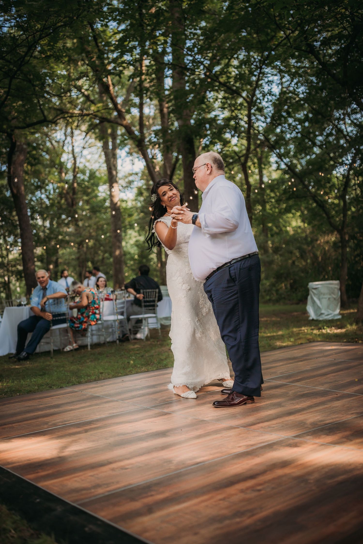 Newlyweds share their first dance on a rustic outdoor dance floor surrounded by trees and watching guests.