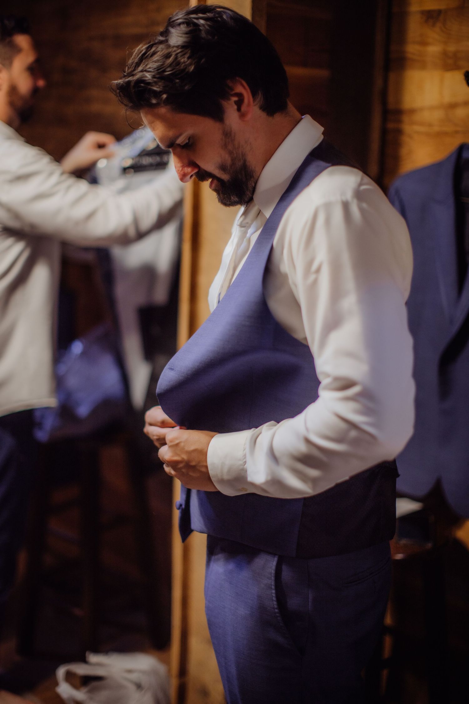 Groomsman adjusting his purple vest while getting ready in a rustic wooden room.