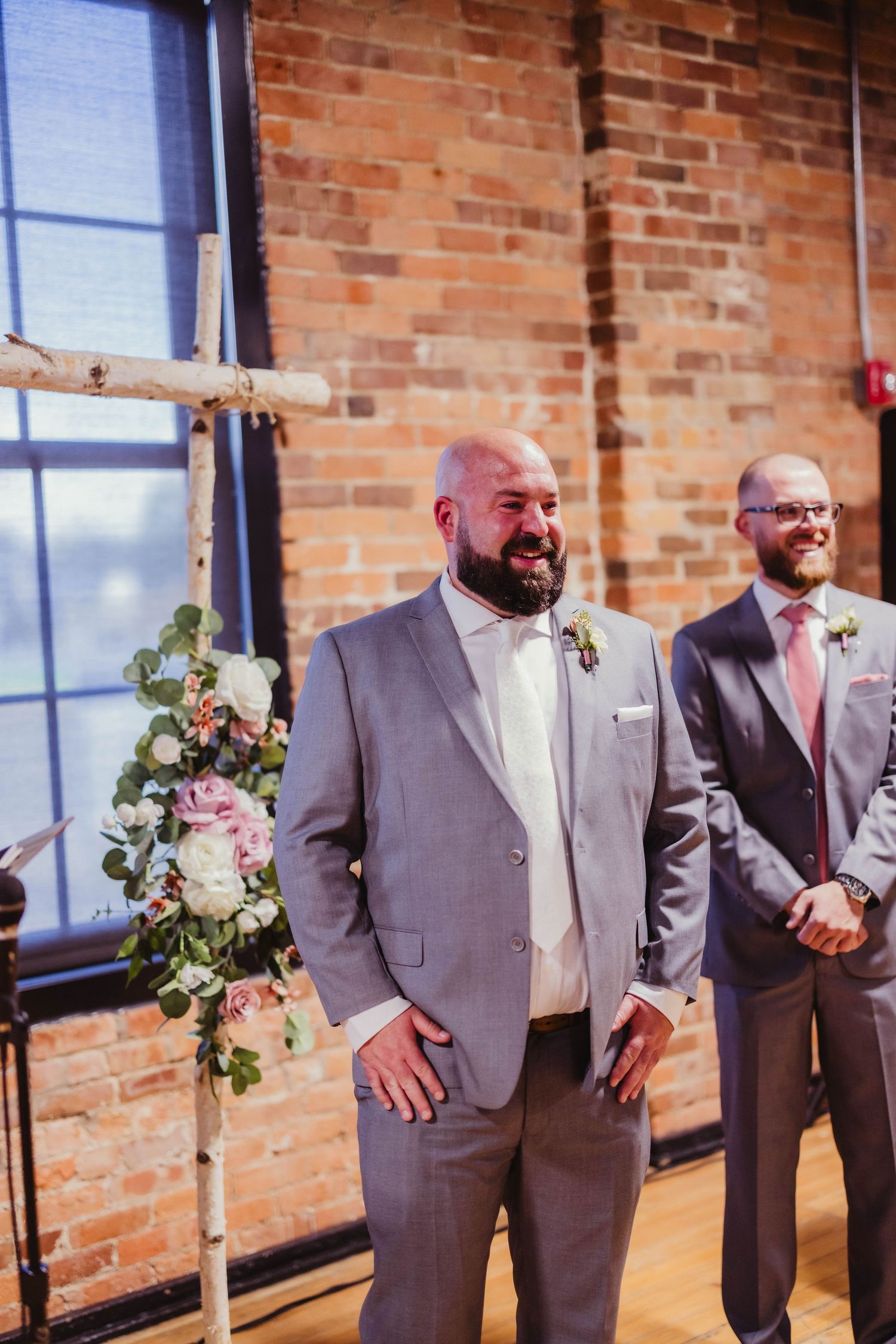 Groomsmen in gray suits stand in front of exposed brick wall during wedding ceremony.