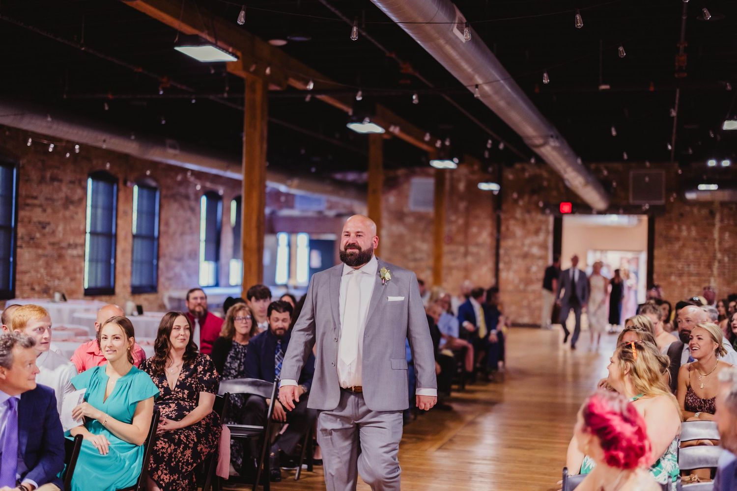 A wedding ceremony participant walks down the aisle of a rustic brick venue with exposed wooden beams and pipes.